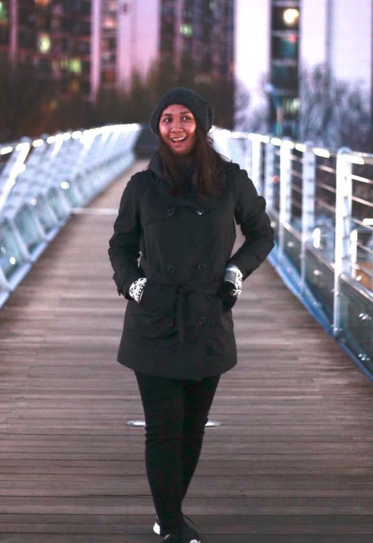 A woman standing on a bridge at dusk, smiling, wearing a black coat, black pants, black hat, and gloves, with city lights and buildings in the background.