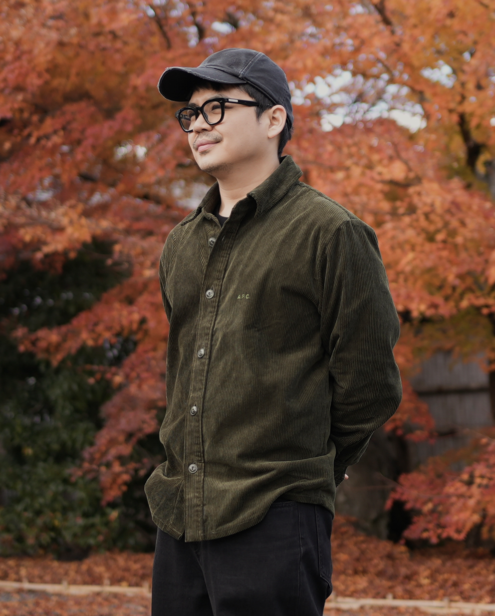 A man with glasses and a baseball cap stands in front of a tree with orange autumn leaves, looking to the side.