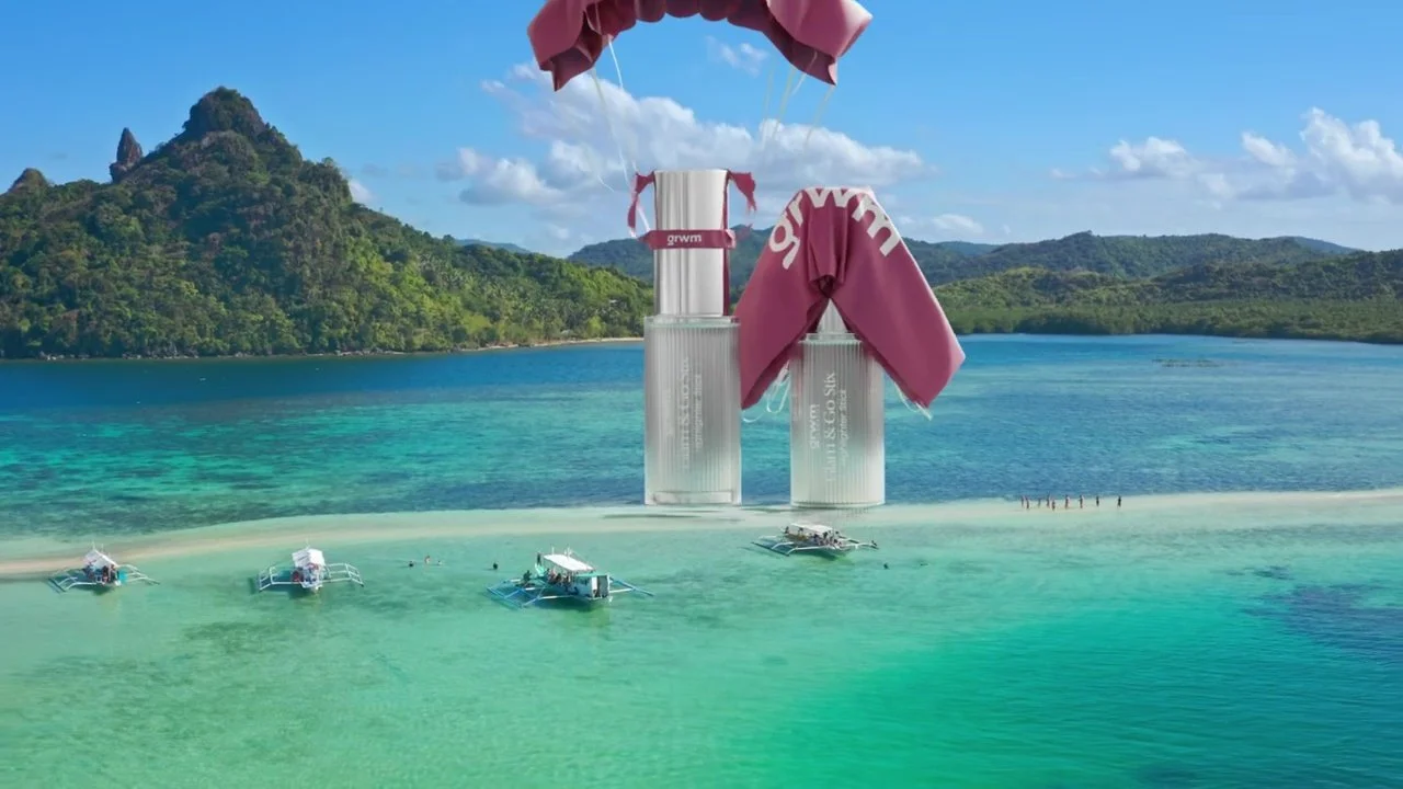 A tropical beach with turquoise water, boats, and lush green mountains in the background. Large perfume bottles are edited to appear as part of the beach scene, with fabric draped over them.