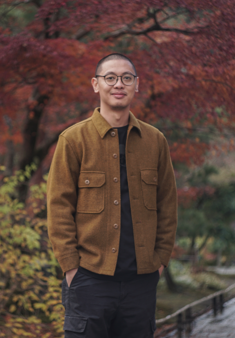 A man with glasses and short hair, wearing a brown jacket and black pants, standing outdoors with autumn foliage in the background.