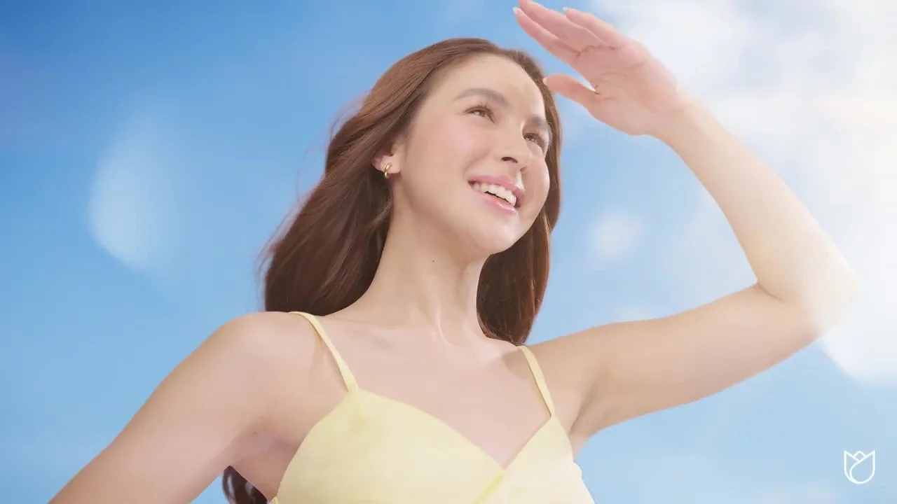 A woman with red hair wearing a yellow top, smiling and shielding her eyes from the sun with her hand against a bright blue sky.