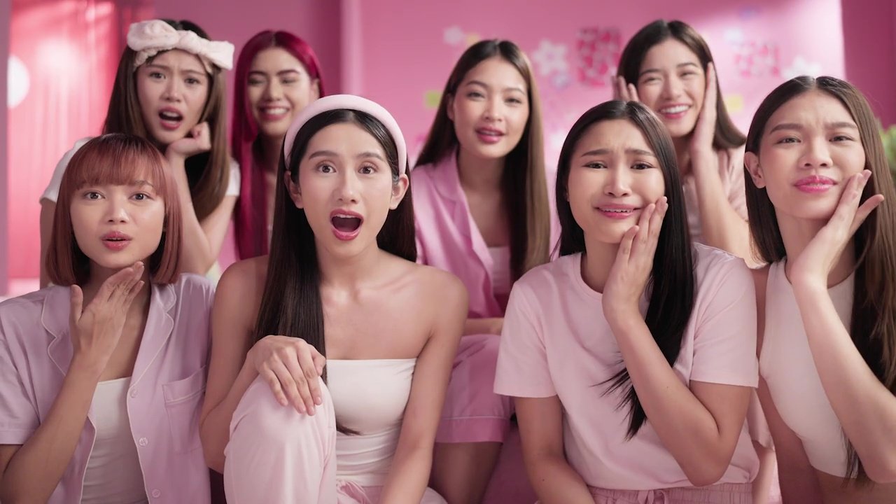 Seven young women wearing pink and pastel pajamas, showing expressions of surprise, concern, and distress, in a pink-themed room.