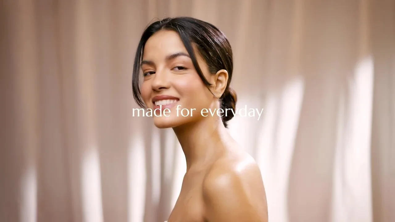 A woman with dark hair styled in a bun, smiling with her head slightly turned to the side in front of beige curtains; the text "made for everyday" is overlaid.