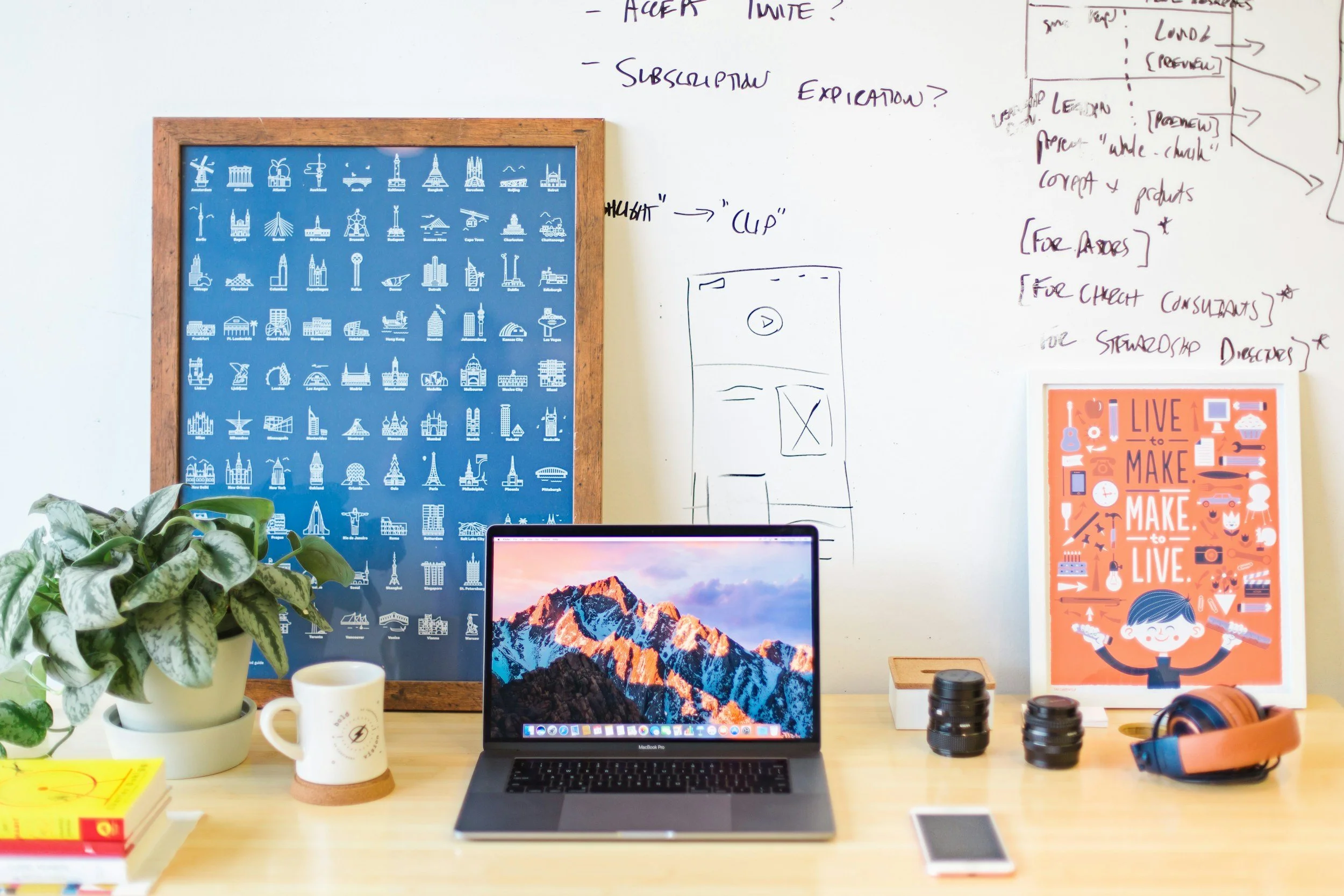 A workspace with a wooden desk holding a laptop, a potted plant, a mug, a smartphone, two camera lenses, and a pair of headphones. Behind the desk, there are two framed posters and a whiteboard with handwritten notes.