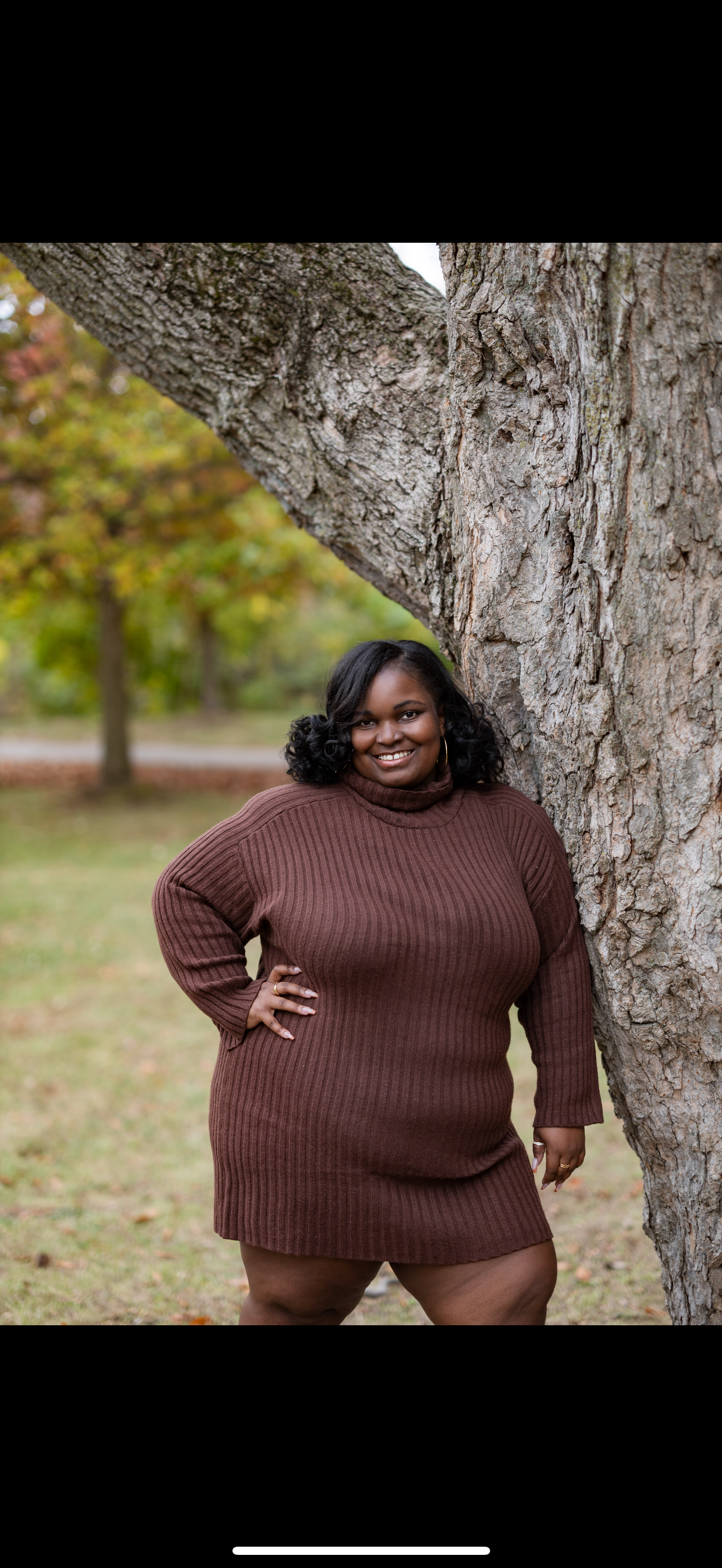 A woman in a brown ribbed sweater dress standing outdoors near a large tree with autumn leaves in the background.