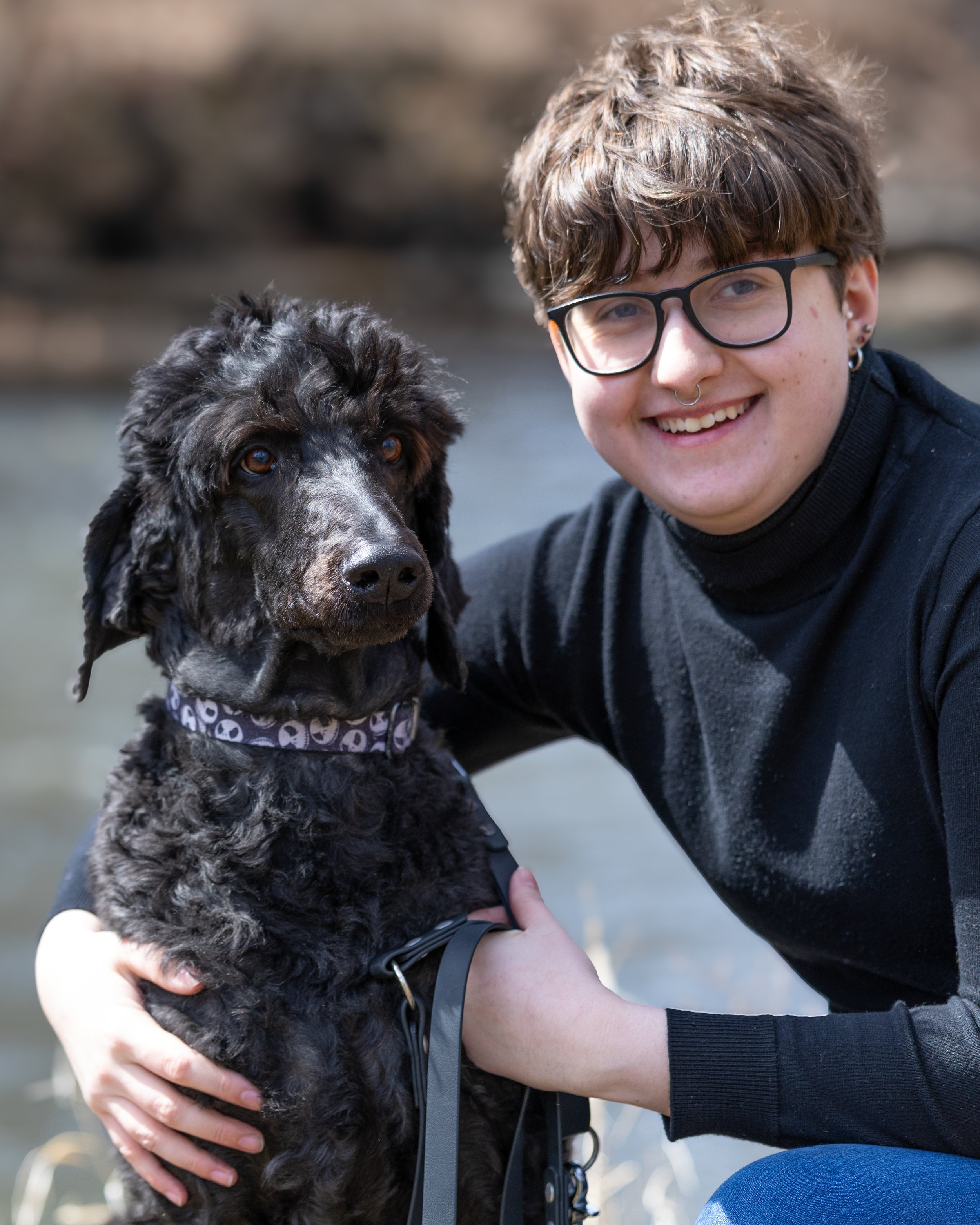 Person in dark sweater with glasses and septum piercing smiling while holding a black and gray puppy wearing a skull-patterned bandana