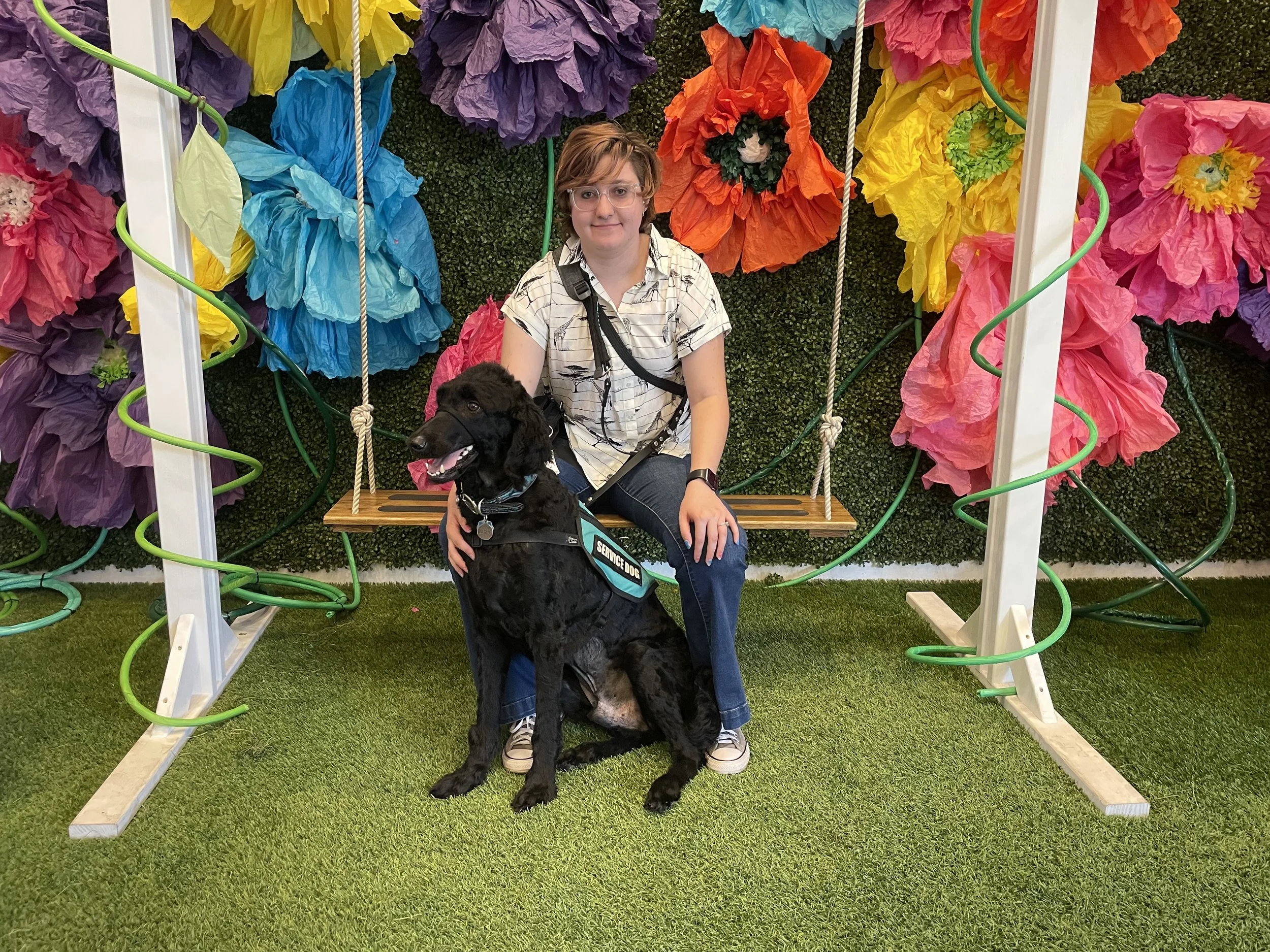 A young person sitting on a wooden swing with a black service dog, in front of a colorful backdrop of large paper flowers in purple, blue, orange, yellow, pink, and green. The person is wearing glasses, a patterned shirt with jeans