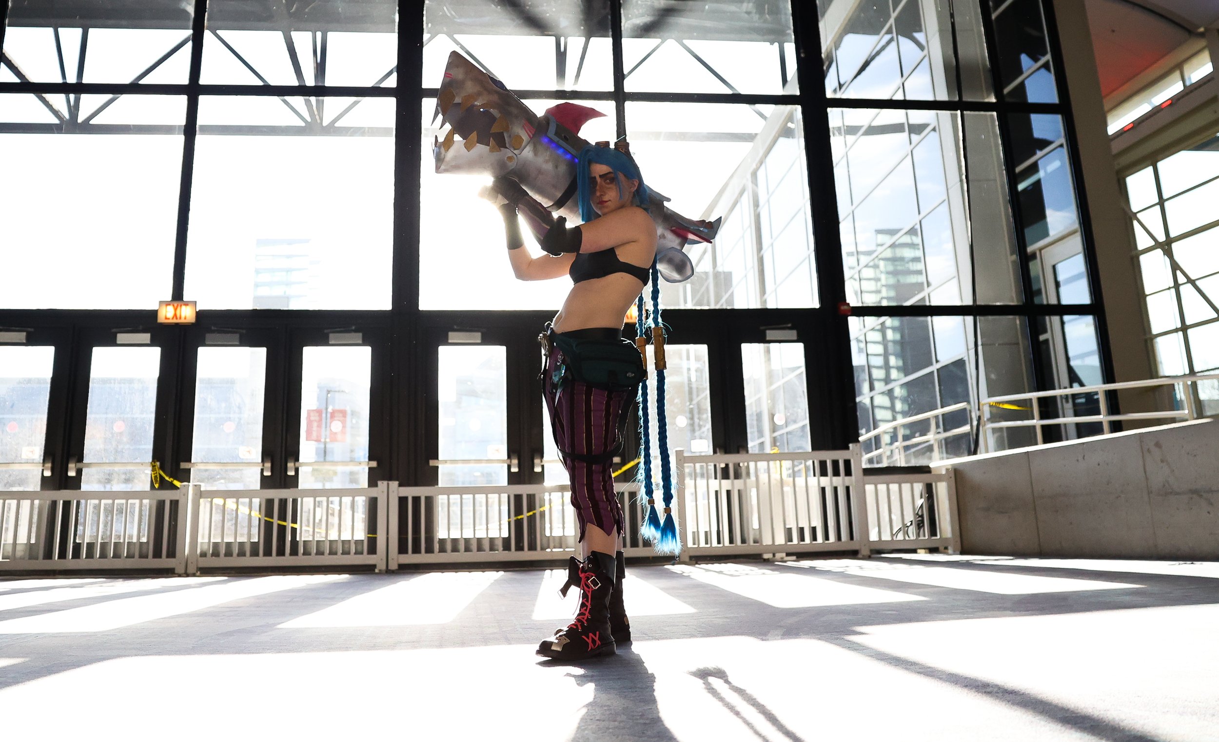 A person with blue hair and cosplay costume holding a large, sword-like prop at a convention center with large windows and a high ceiling.