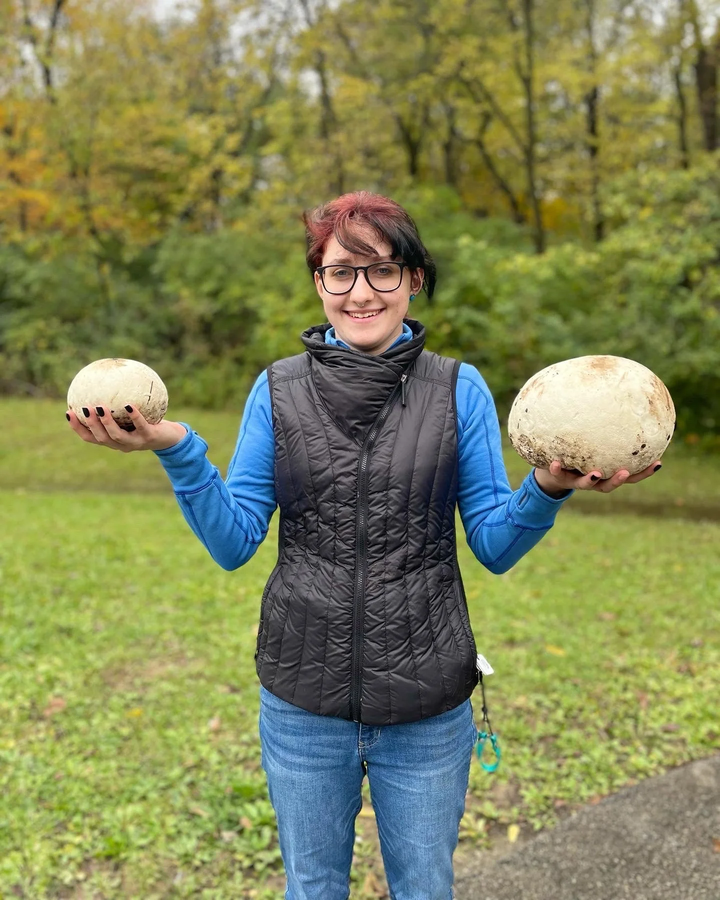A person with short hair and glasses holding two large, round mushrooms outdoors with a background of trees with yellow and green fall foliage.