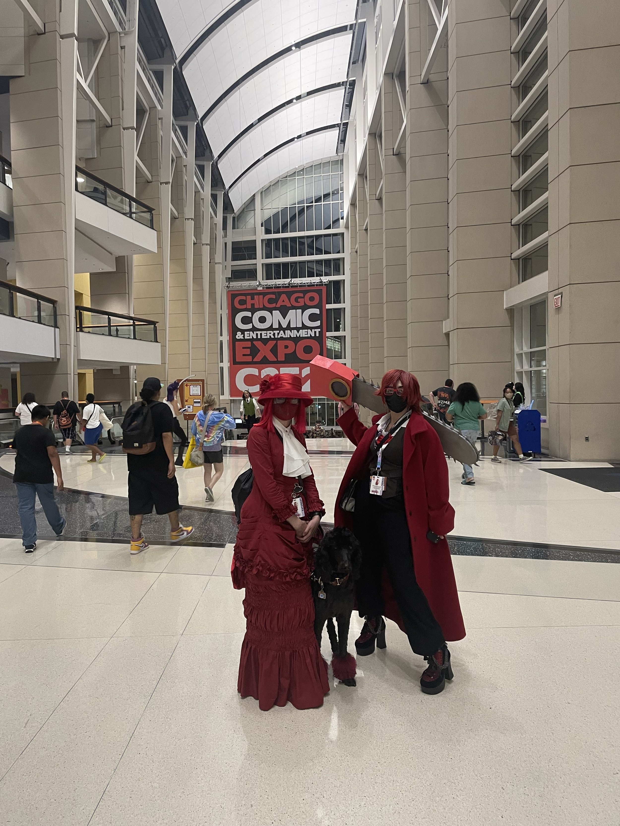 Two individuals in cosplay costumes posing for a photo at the Chicago Comic & Entertainment Expo (C2E2) inside a large, modern convention center with high arched ceilings and a large red and black sign in the background.