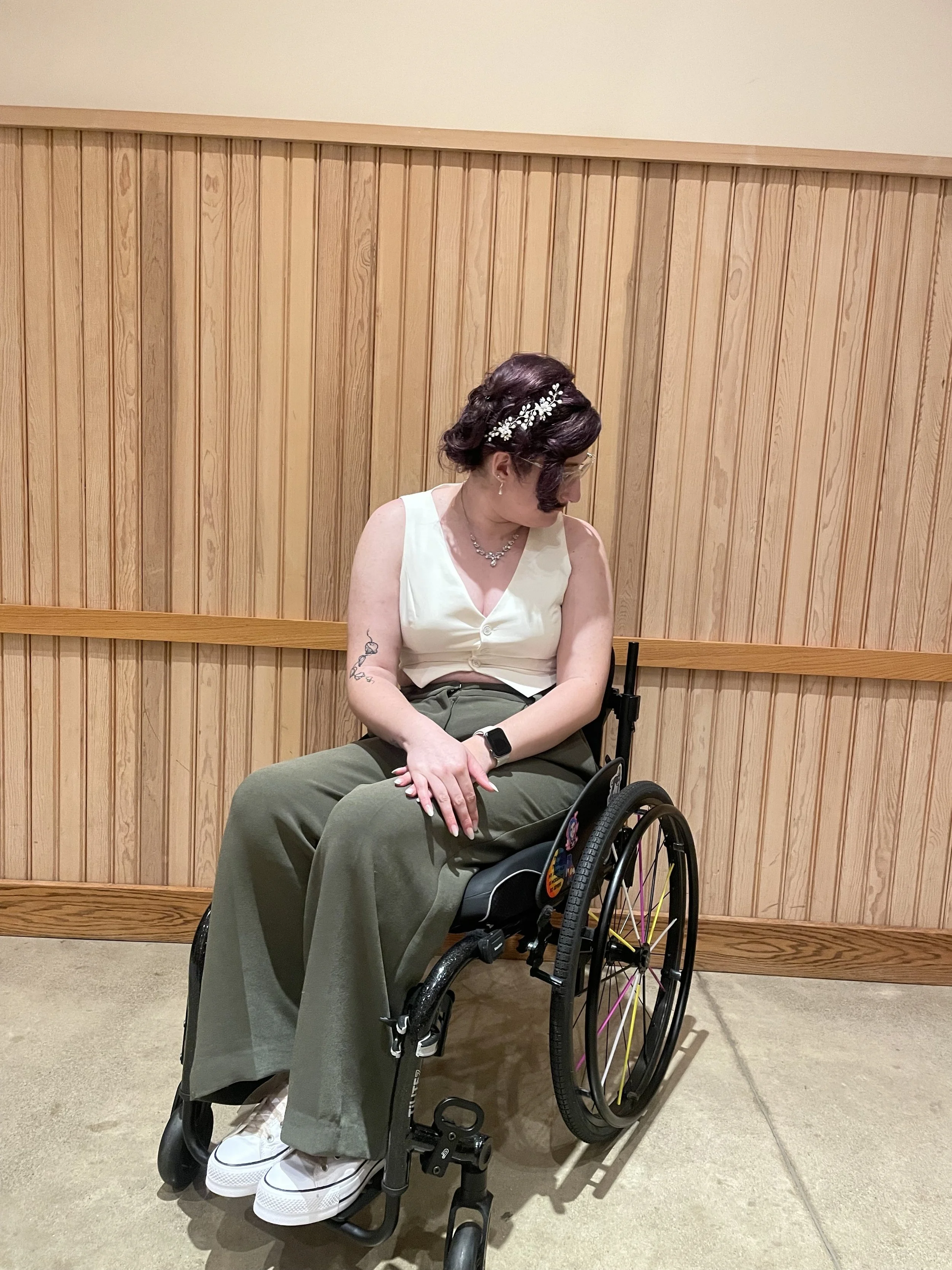 A person with dark hair styled in an updo with a floral headband, sitting in a wheelchair with tattoo on their arm, wearing a sleeveless cream top, olive green pants, white sneakers, looking down, against a wooden wall background.