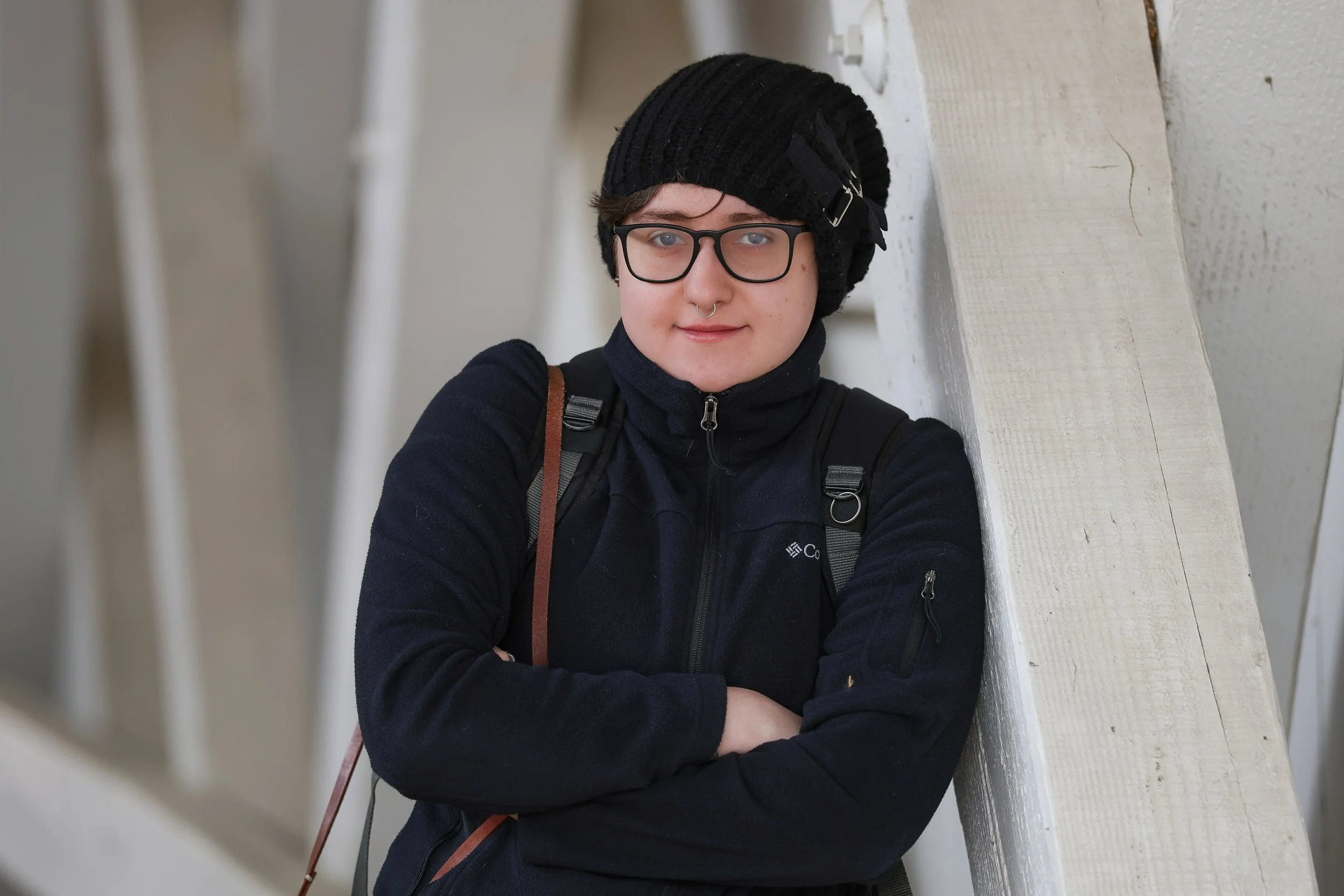 A young person with glasses, a septum piercing, wearing a black beanie with a bow, a dark jacket, and a backpack, leaning against a white wall with a neutral background.