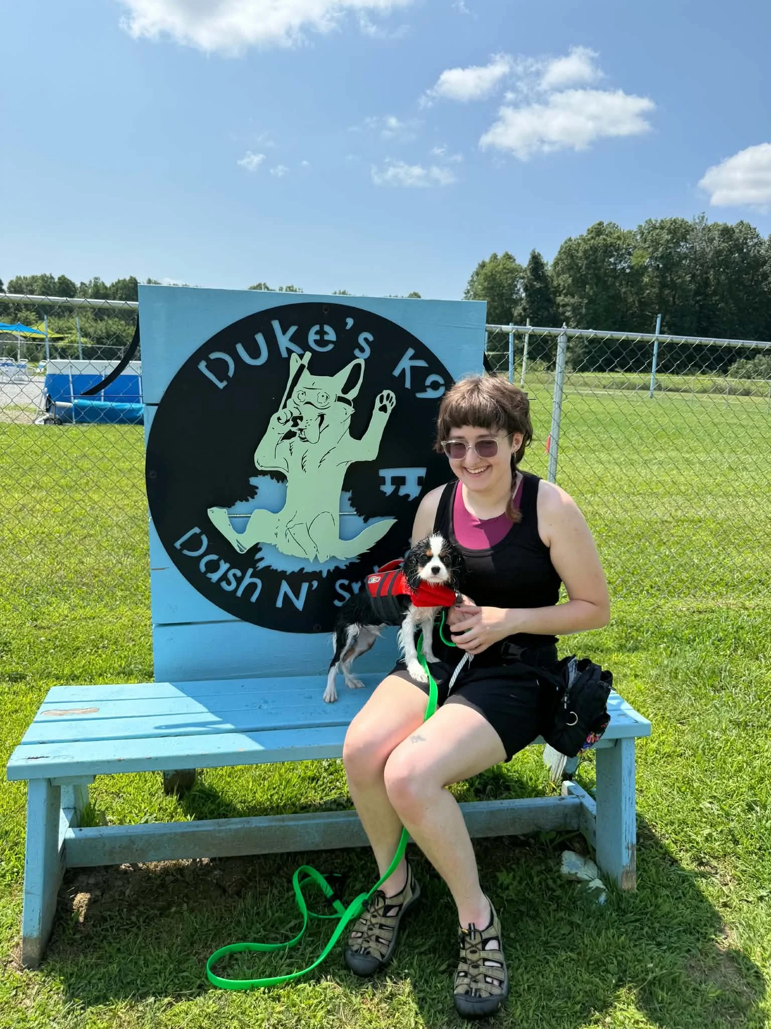A woman with sunglasses and a sleeveless black tank top sitting on a light blue park bench, holding a black and white puppy dressed in a red outfit. A large blue sign with a cartoon dog and the text 'Duke's K9' is behind her. Green grass and trees ar