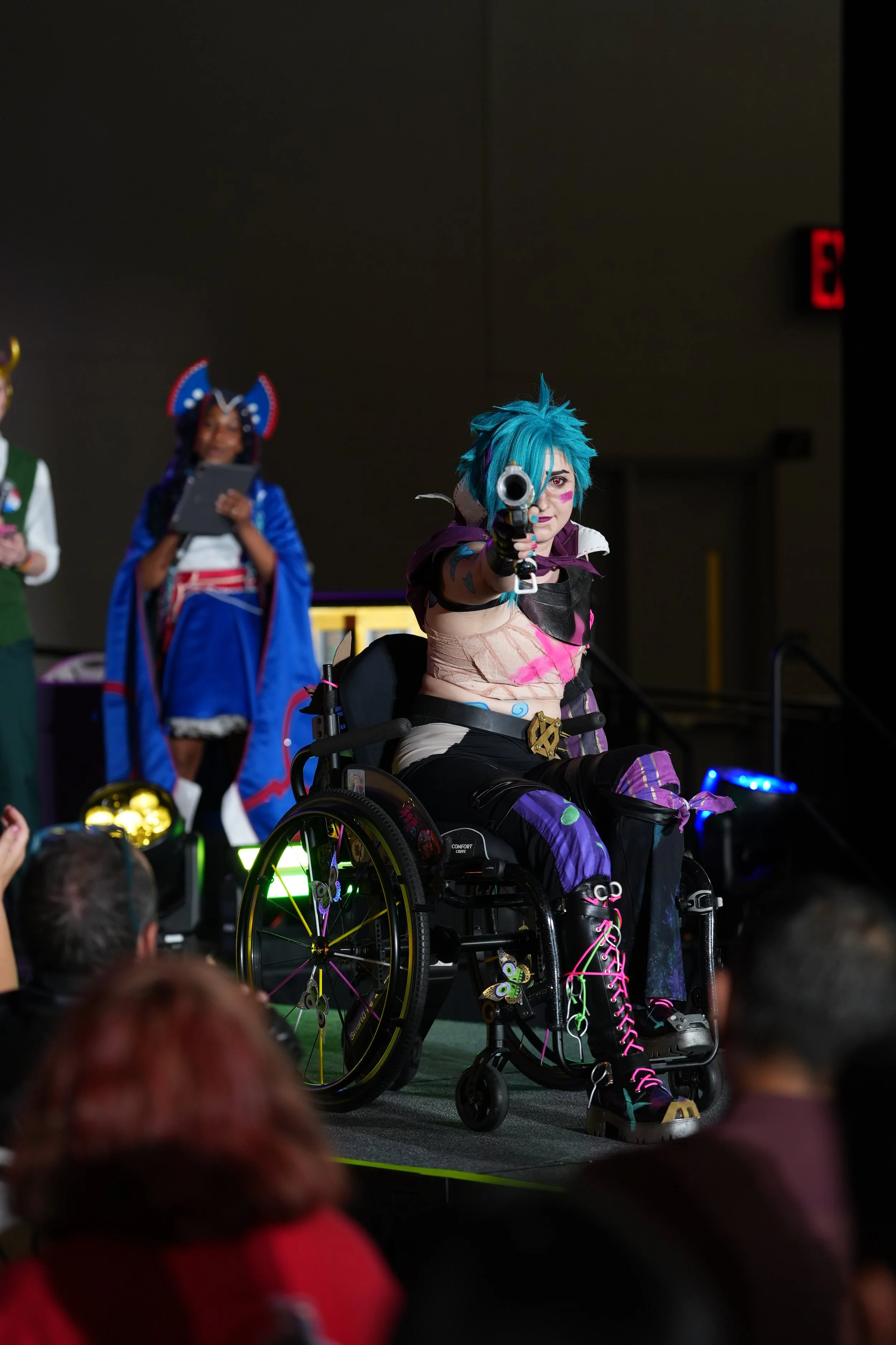 Person with bright blue hair in a wheelchair pointing a gun on stage during a cosplay event, with other cosplayers in the background.