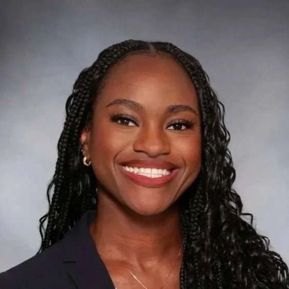 A woman with long, curly black hair, smiling, wearing a dark blazer and a white blouse, with gold earrings and a necklace, against a neutral gray background.
