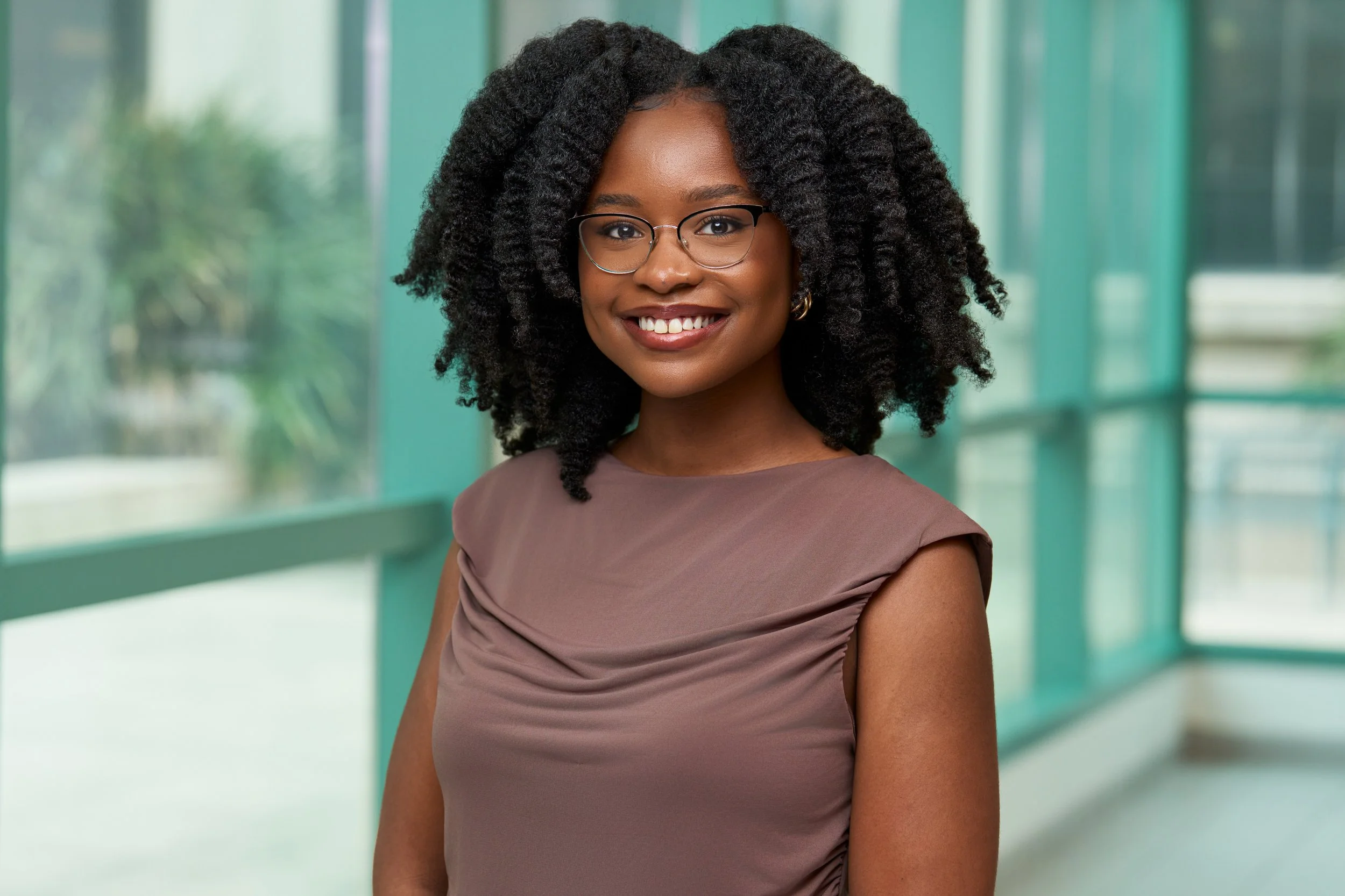 Portrait of a young Black woman with braided hair, smiling, wearing a gray blazer, white shirt, and pearl earrings, against a gray background.
