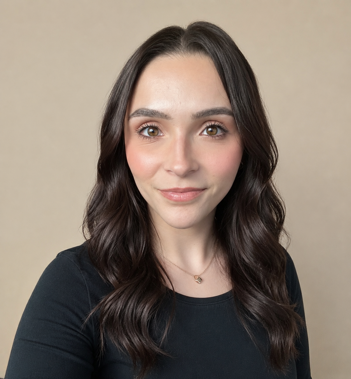 A young woman with long wavy dark brown hair and hazel eyes, smiling slightly, wearing a black top and a delicate gold necklace, posing against a beige background.