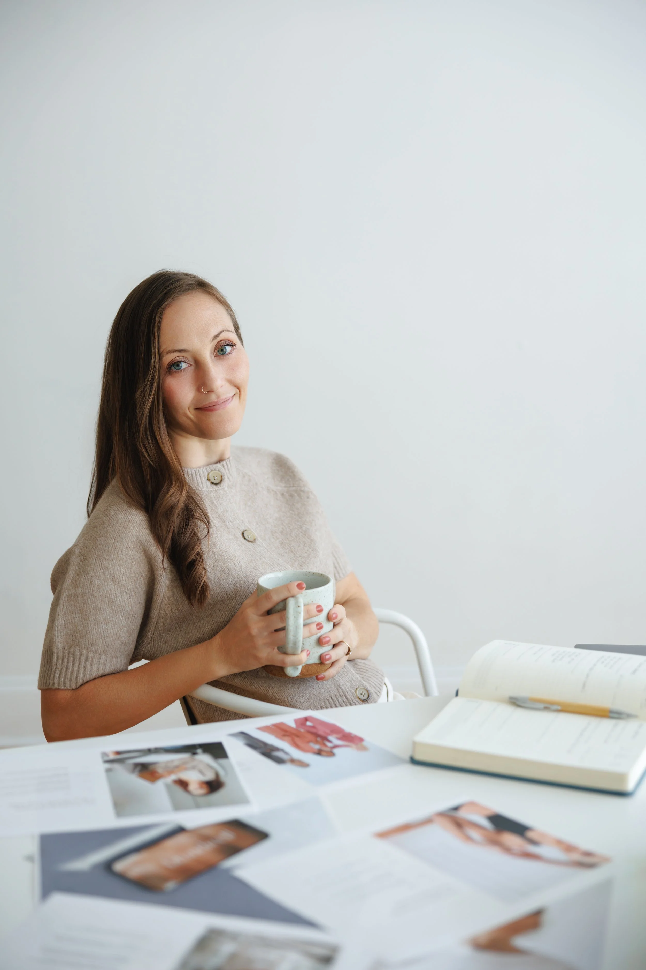 A woman with long brown hair and blue eyes sitting at a table with various papers, magazines, and a book, holding a mug in her hands, smiling gently at the camera, in a bright minimalist room.