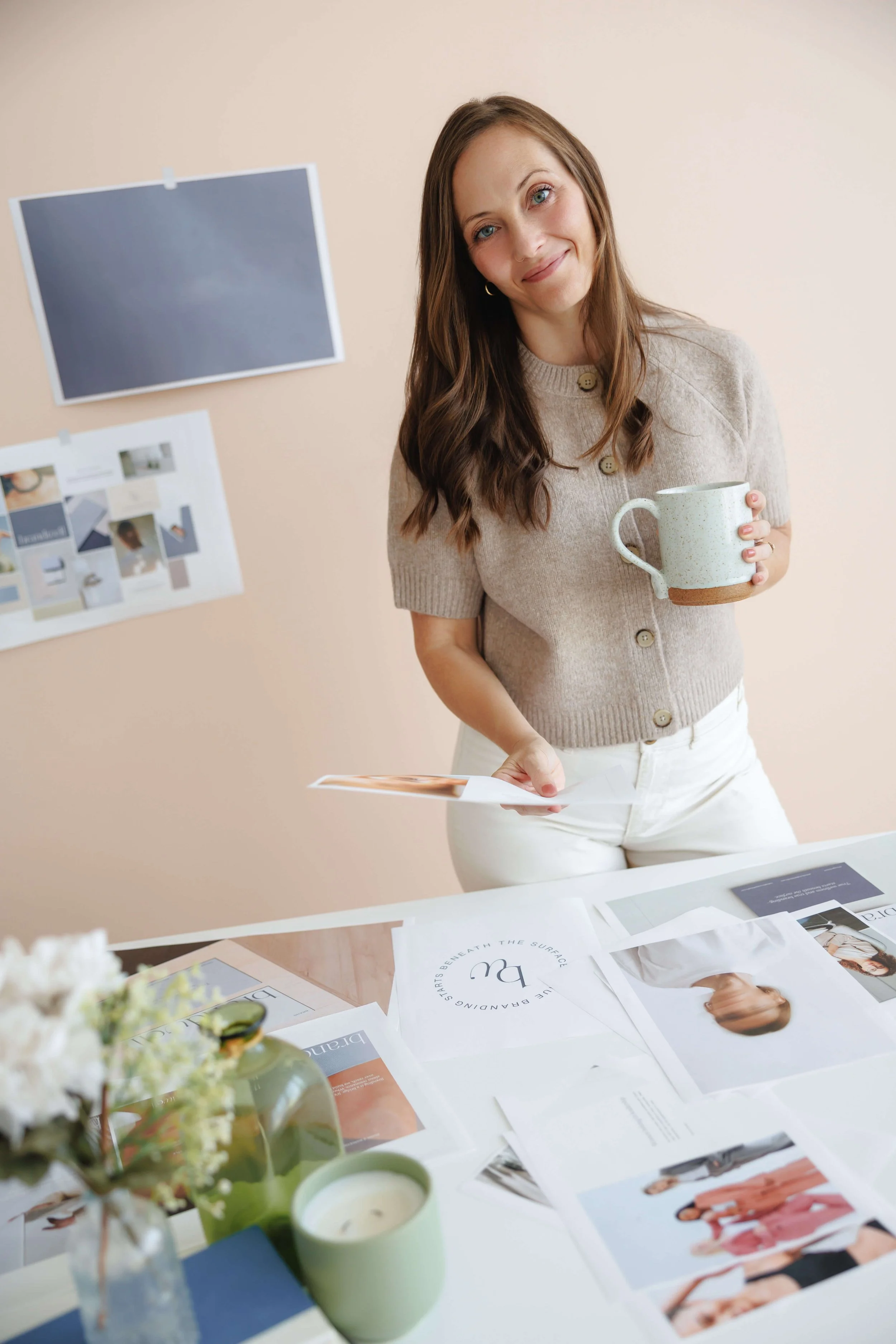 A woman with long brown hair, wearing a beige button-up shirt, holding a white ceramic mug, smiling at the camera in an office or studio setting with a bulletin board and various papers and magazines on a table.