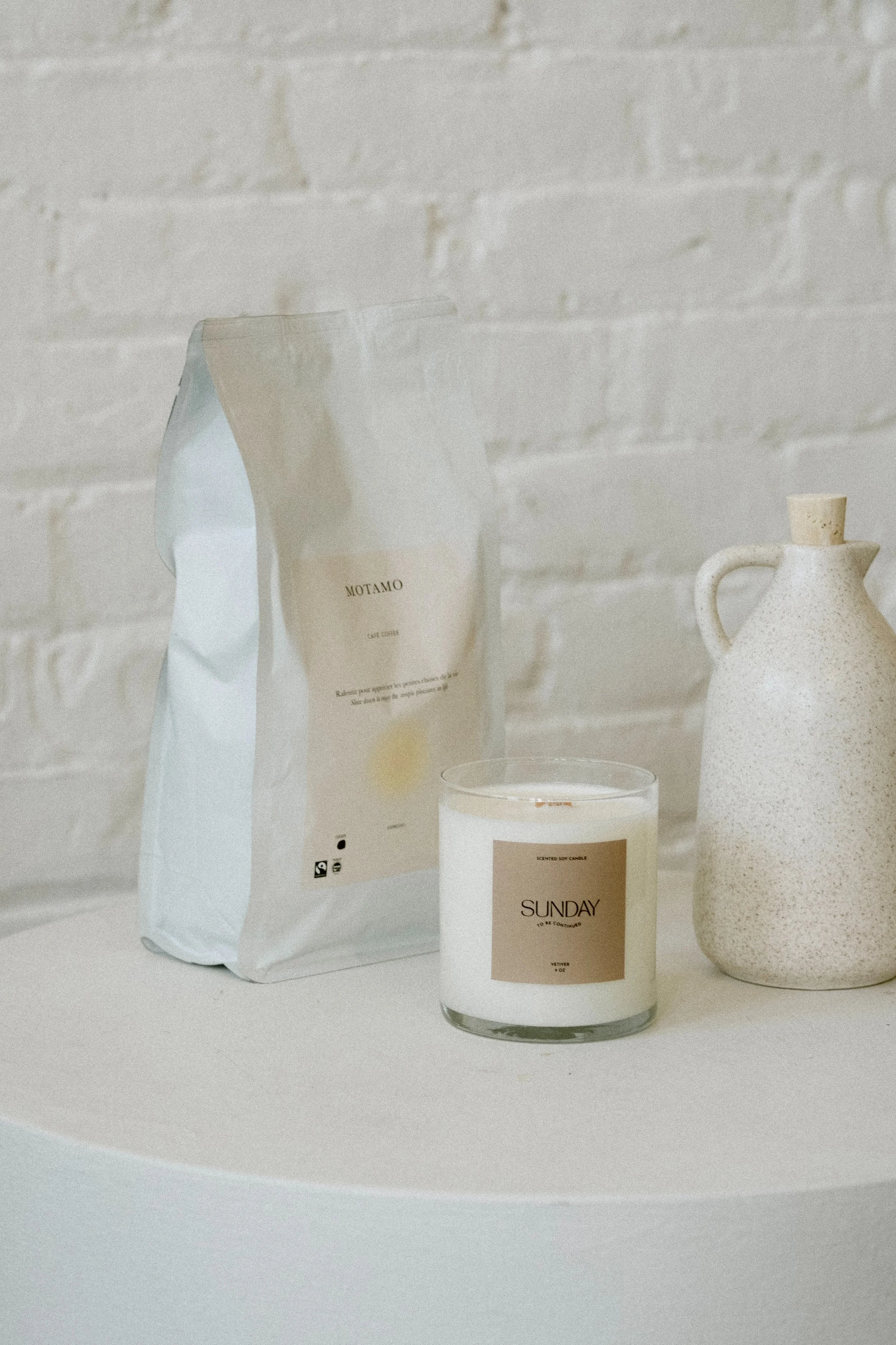 A white brick wall background with a white table holding a white candle labeled "SUNDAY," a ceramic vase, and a white coffee bag.