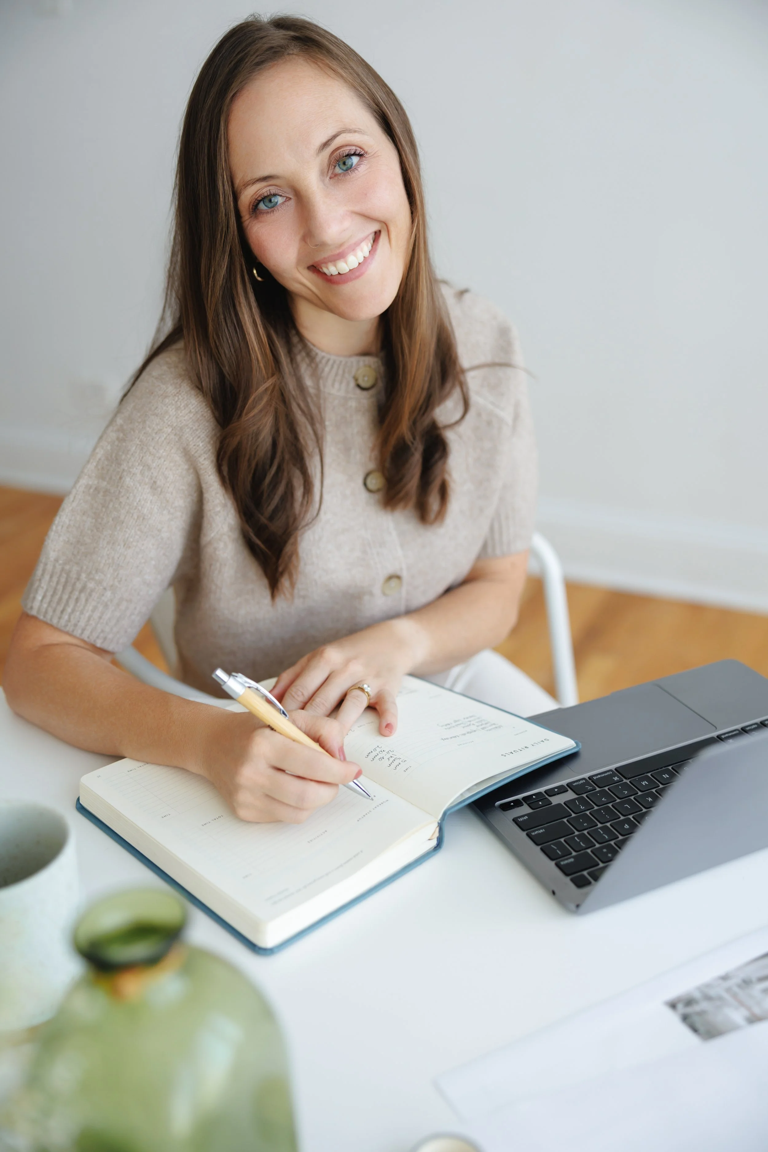 A young woman with blue eyes and long brown hair, wearing a beige button-up top, sitting at a white desk. She is smiling and writing in a notebook with a pen. A laptop is open in front of her, and there are two green avocados in a white bowl on the desk. The background is a plain light-colored wall.