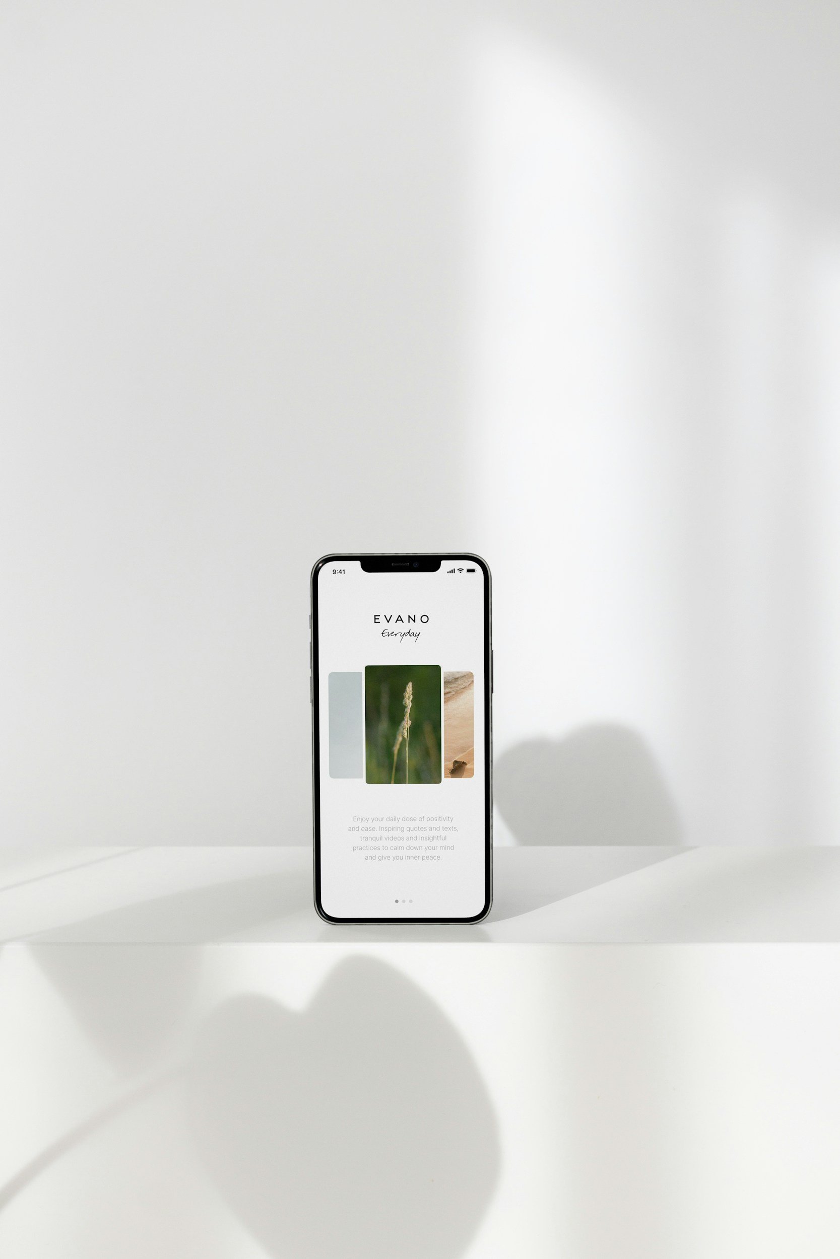 A smartphone displays the Evano app on a white surface with a plain white background. The screen shows the app's interface with images of plants and a text description about positivity and mindfulness.