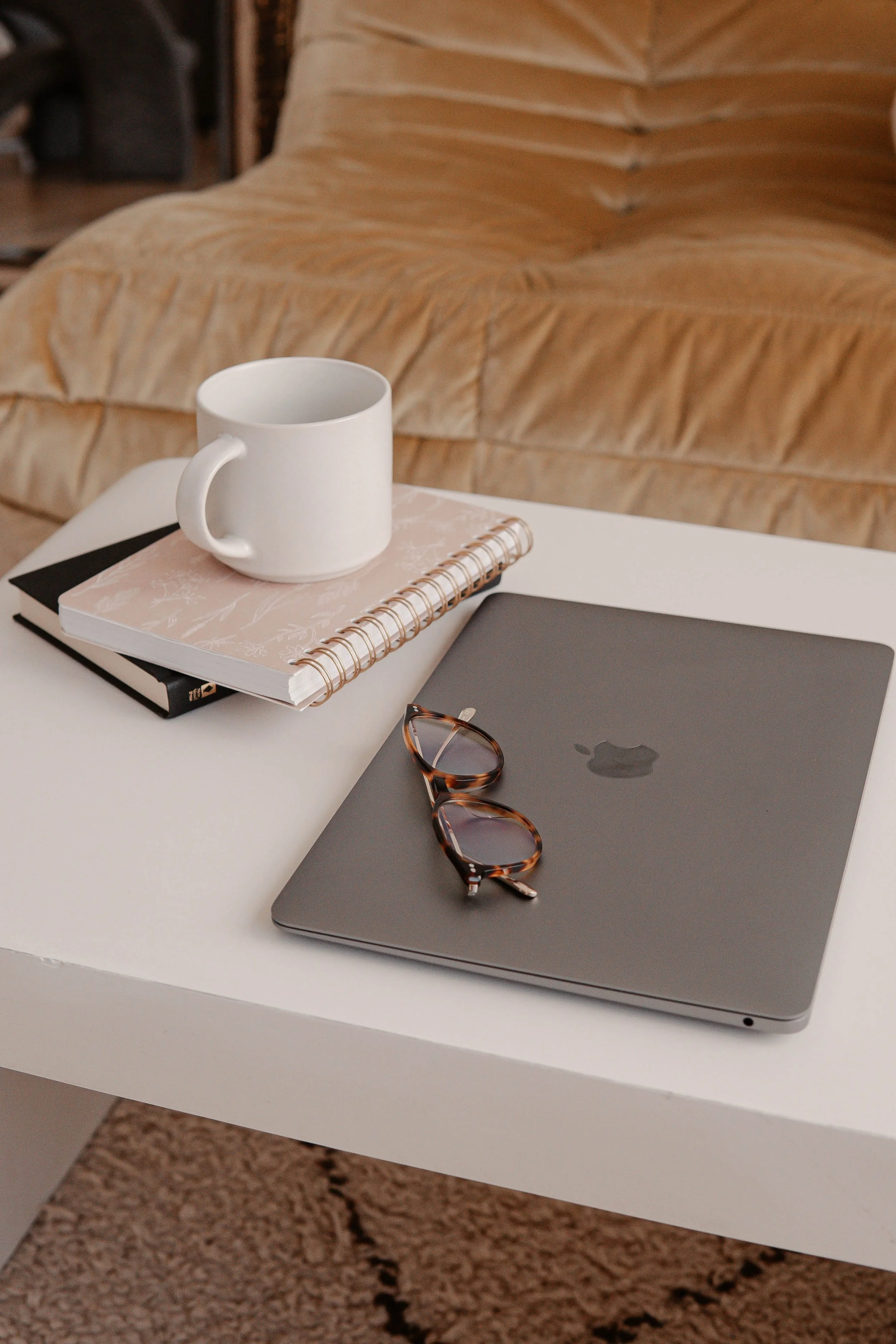 A white mug, a notebook, a silver laptop with glasses on top, and a black notebook are on a white table. In the background, there is a beige upholstered chair.