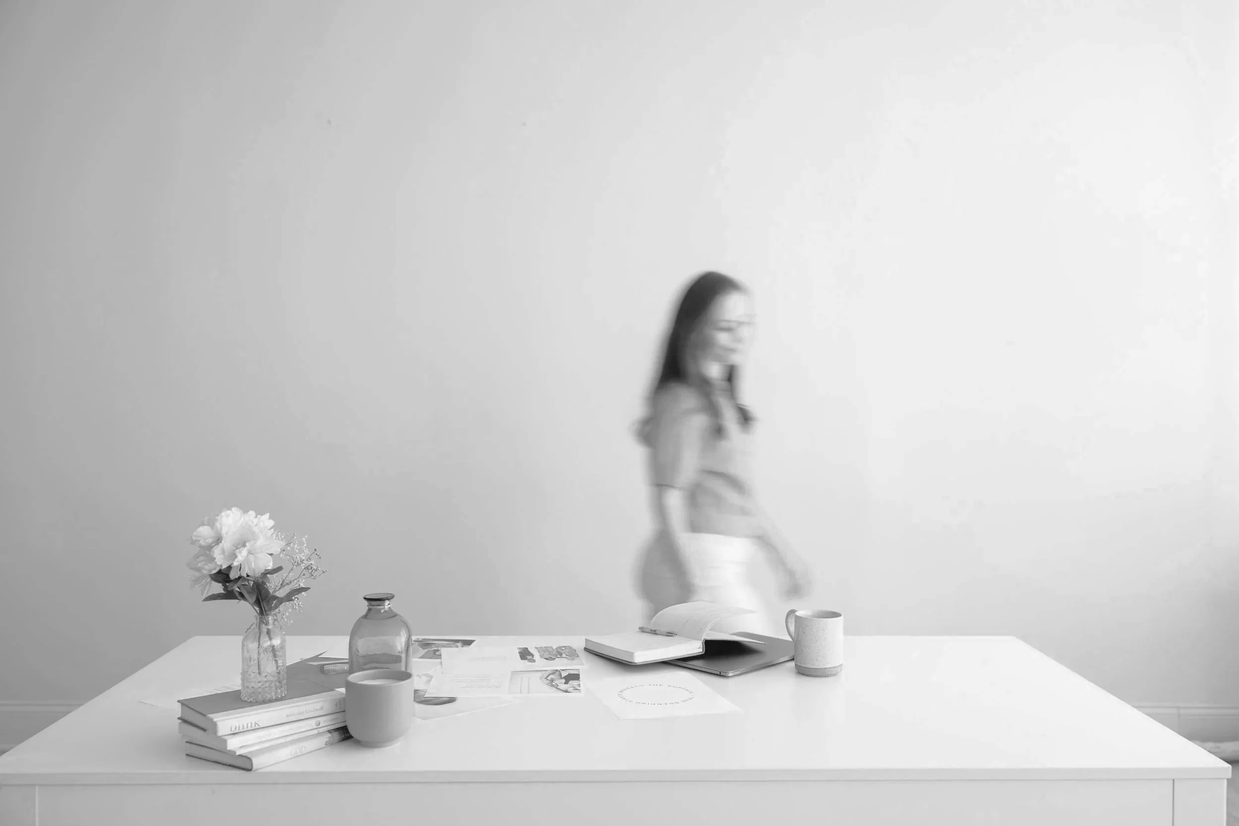 A minimalist black-and-white photograph of a modern workspace. It features a white table with a small collection of books, a glass vase with flowers, three cups, and some papers and magazines. In the background, a person with long hair and glasses stands, holding a phone or small device, against a plain, light-colored wall.
