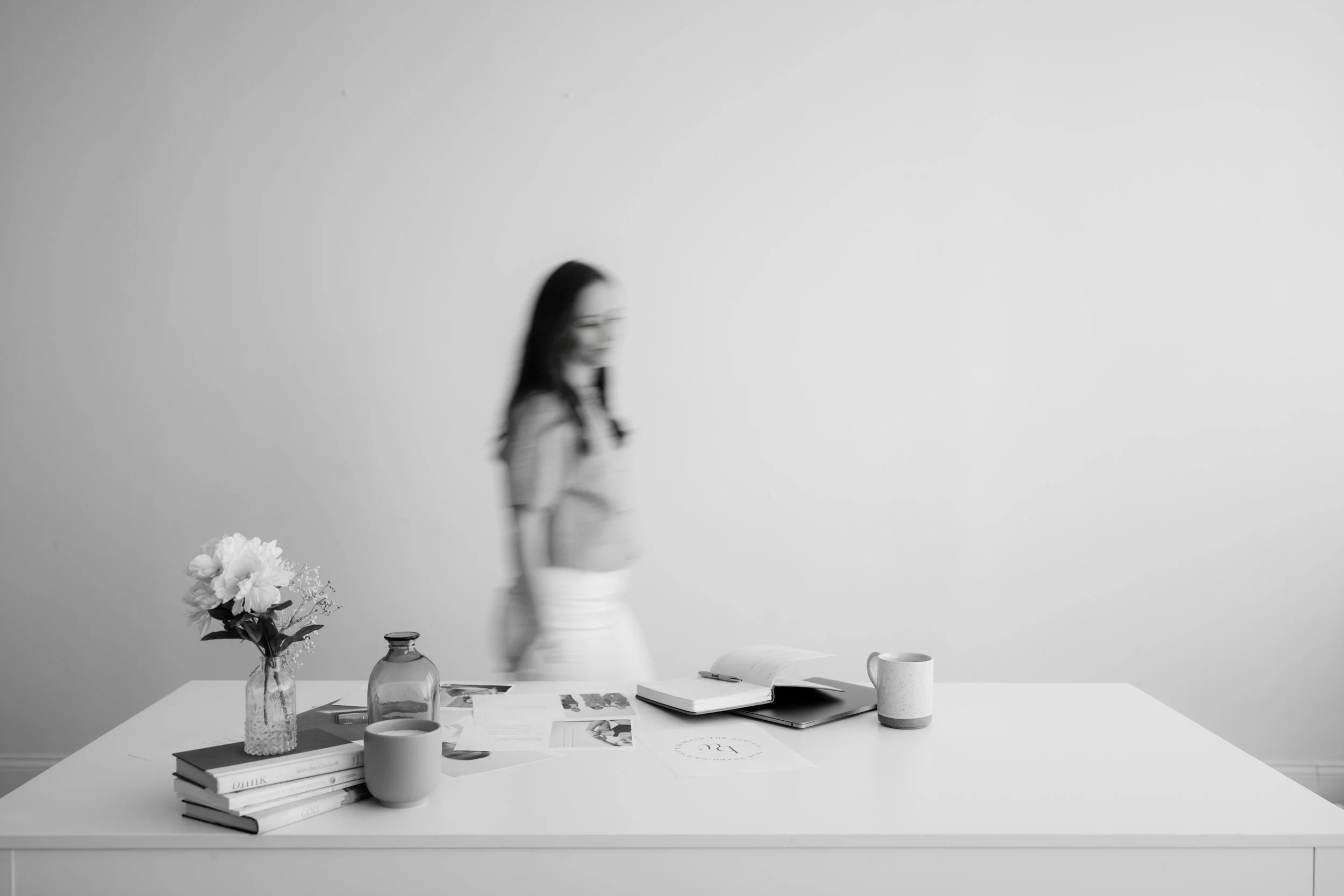 A blurred woman standing behind a white desk with books, candles, a flower vase, open notebook, and coffee mugs, against a plain wall.