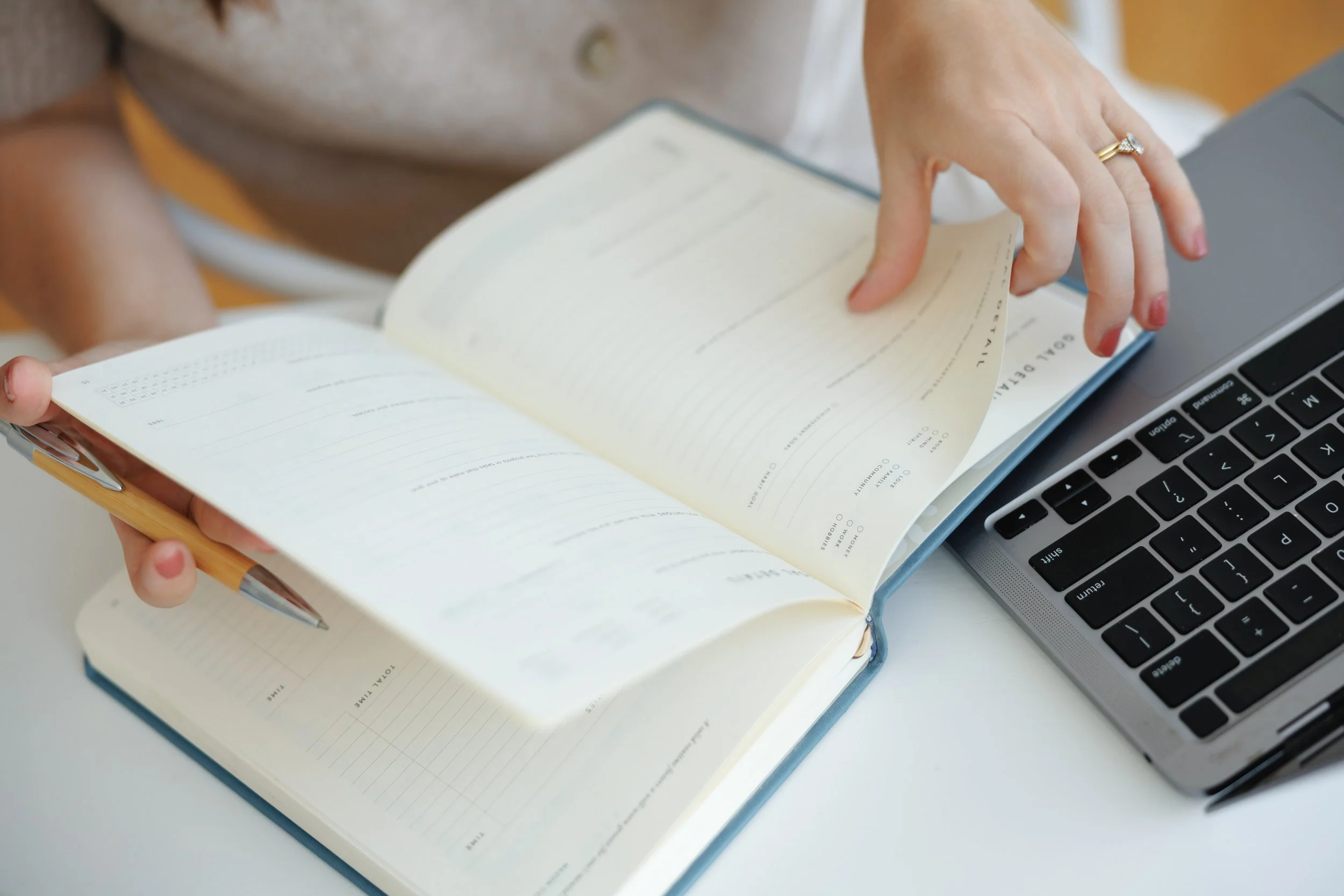 Person flipping through a planner with a pen in hand, next to a laptop on a white desk.