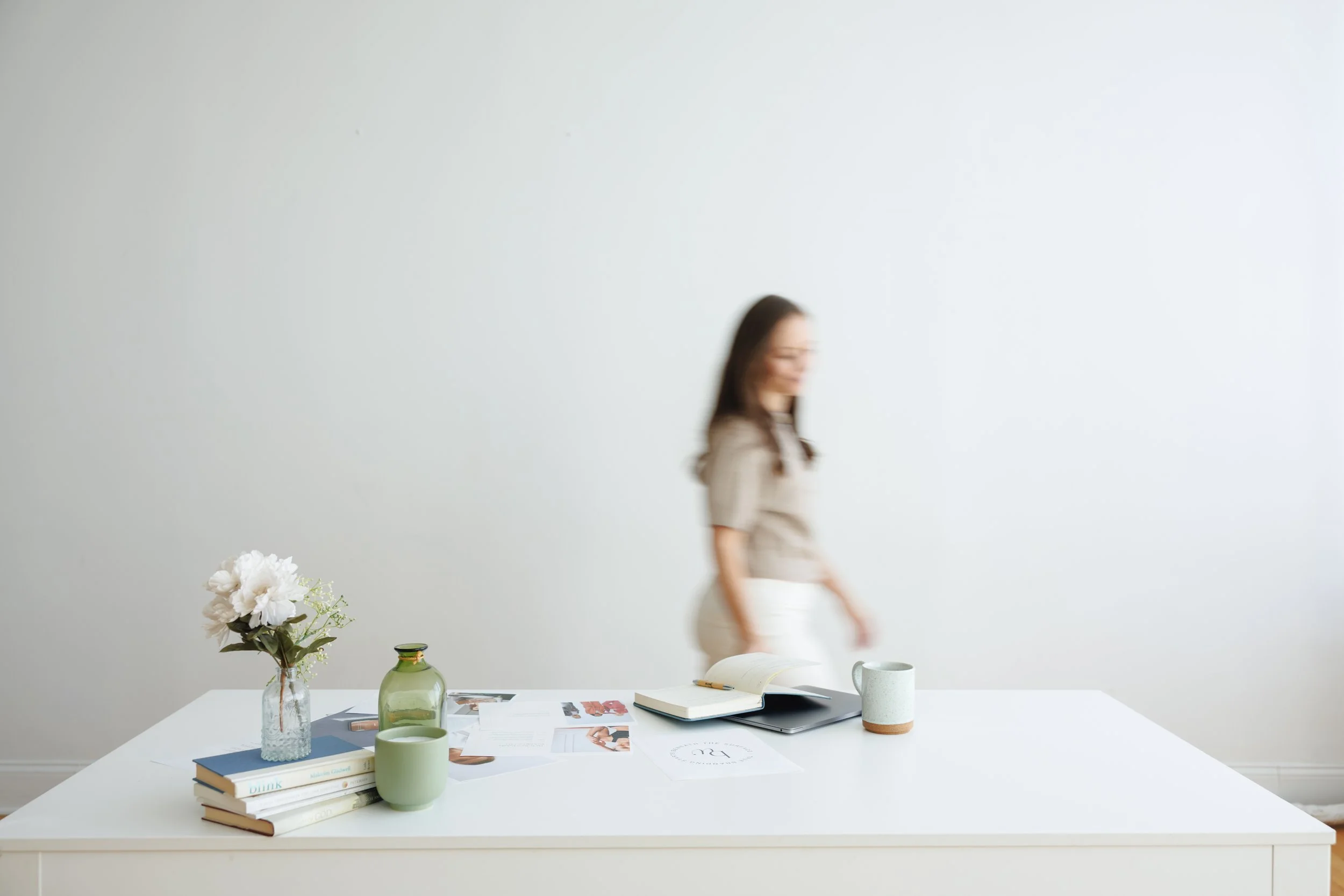 Blurred woman walking past a white table with books, magazines, framed photos, a green vase, and cups on it.