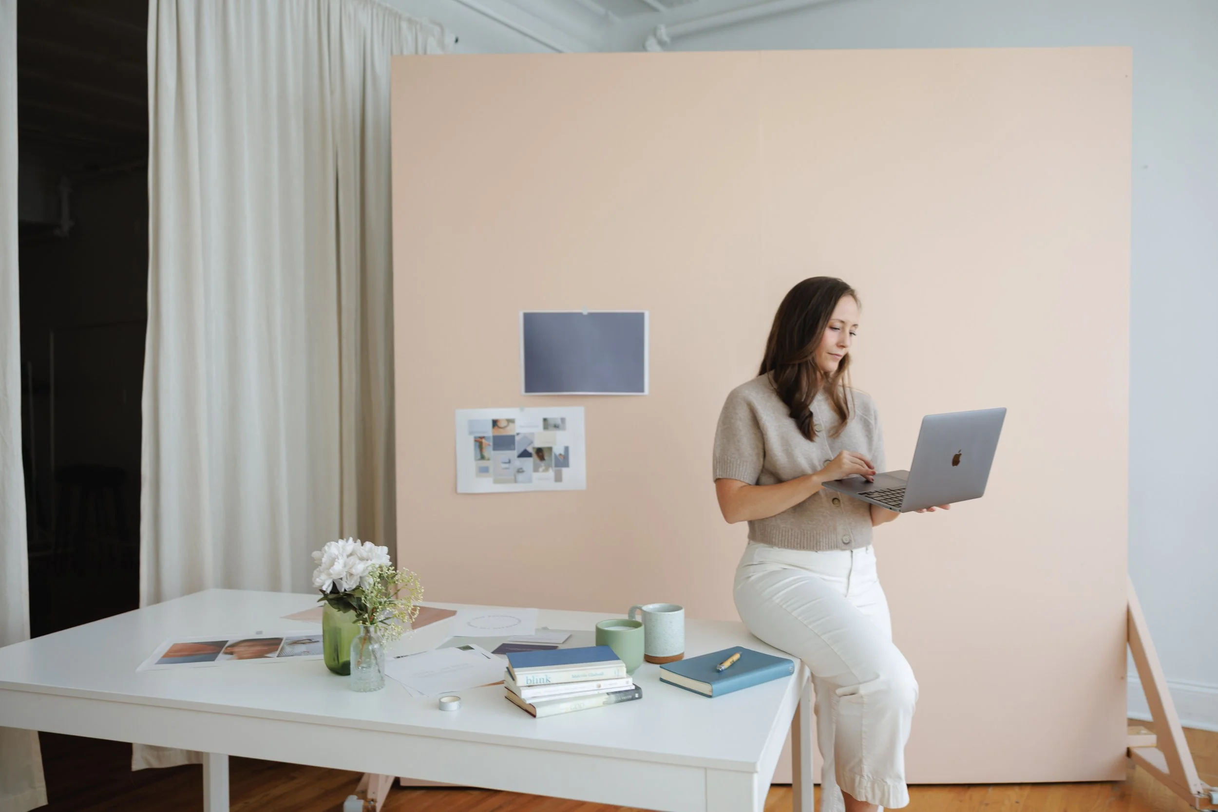 A woman with long brown hair wearing a beige sweater and white pants holds a laptop while standing in a modern office or creative workspace. The space features a white table with various books, papers, a vase with white flowers, and coffee mugs, with a beige partition behind her. White curtains cover part of the background wall.
