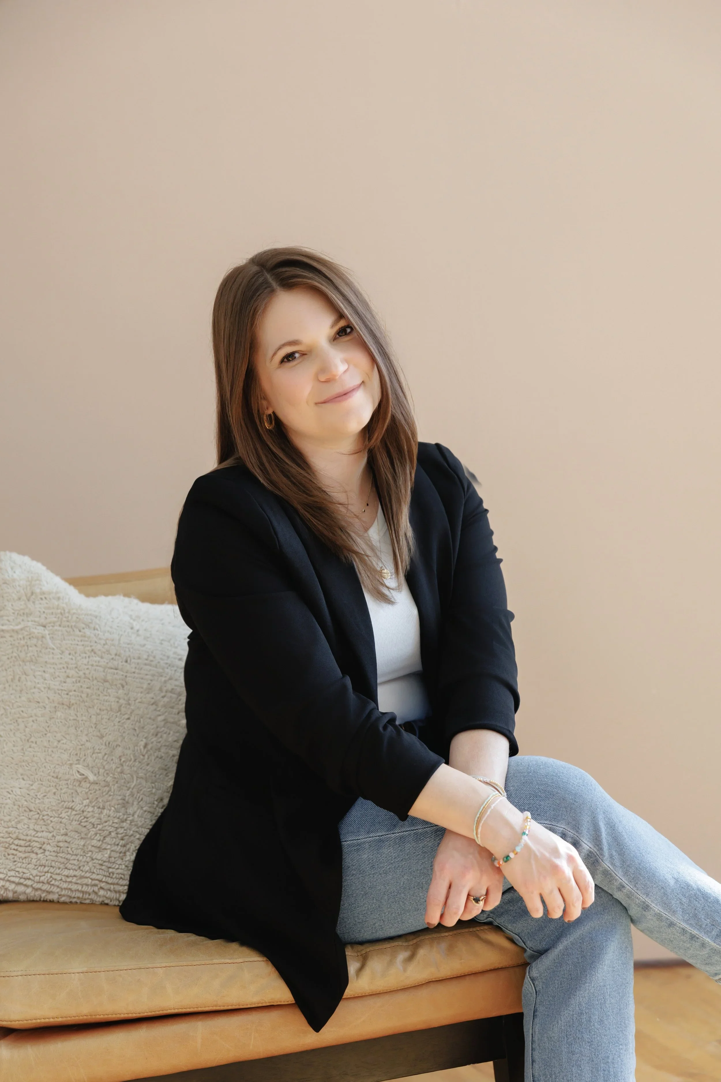 Brown haired woman with black blazer sitting on tan chair with neutral back