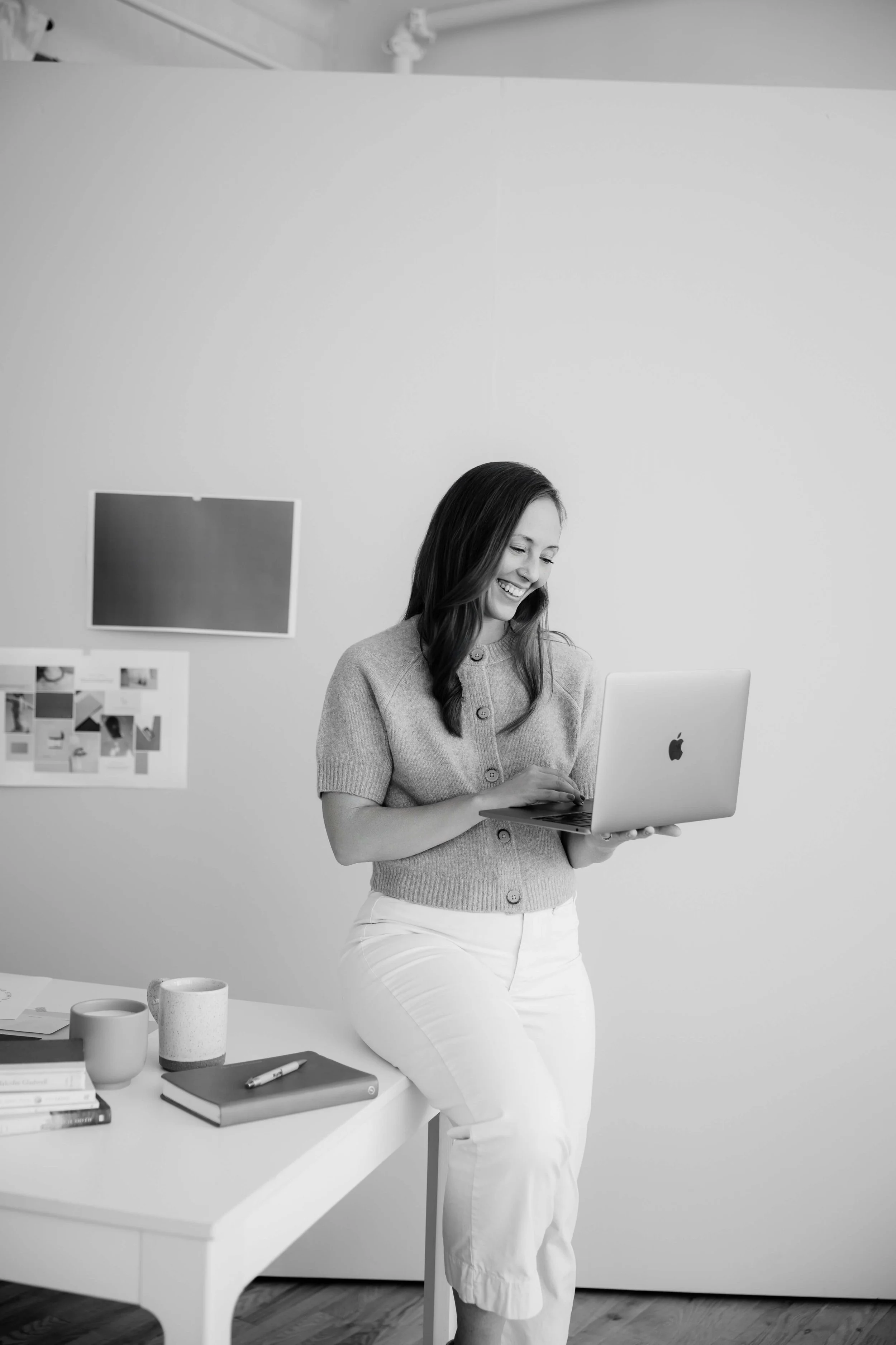 A woman standing in an office, holding a laptop and smiling. Behind her, a white wall with a small bulletin board displaying photos and notes.