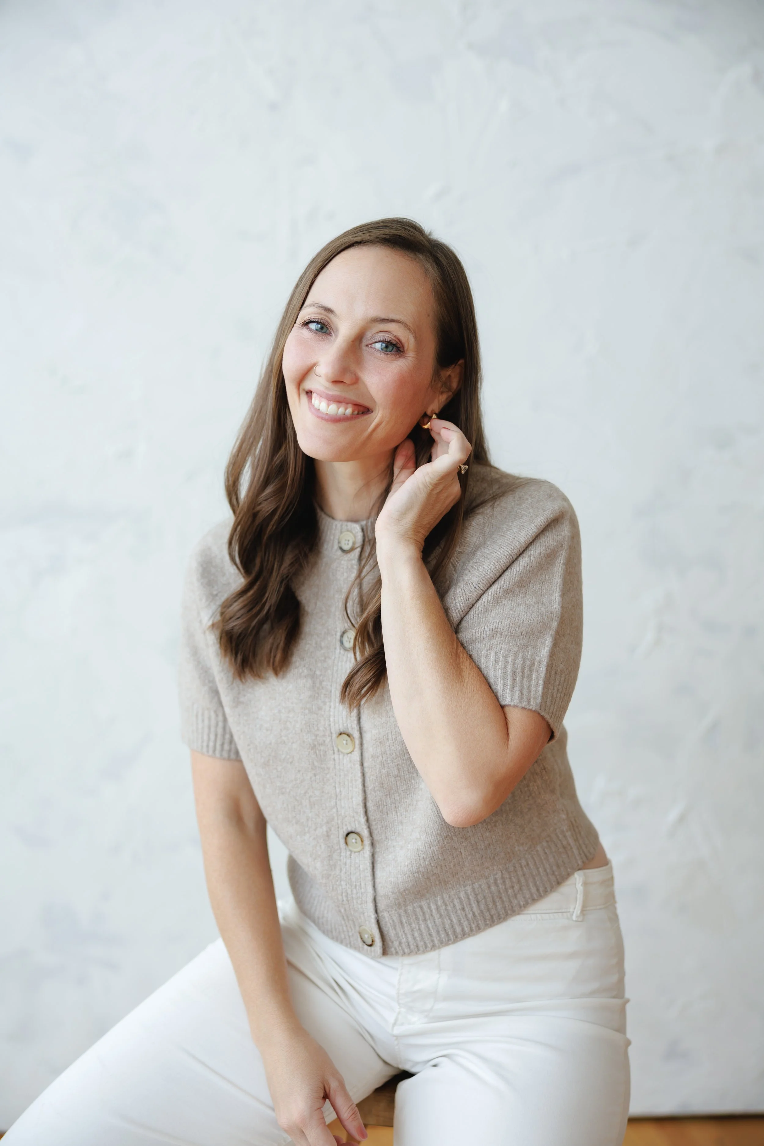 A woman with long brown hair and blue eyes smiling softly, sitting against a white wall, wearing a beige short-sleeved button-up sweater and white pants.