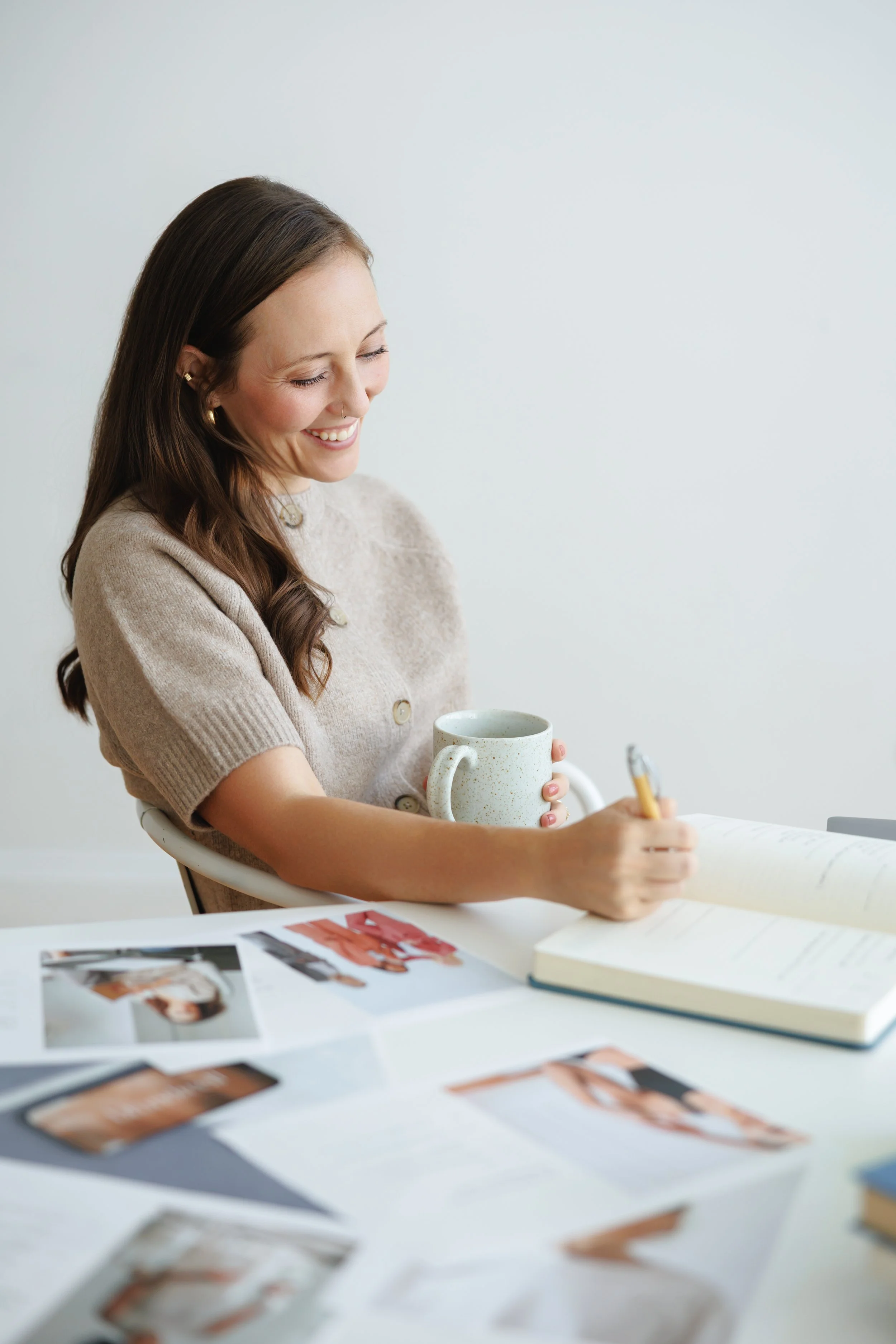 A woman smiling and writing in a notebook while holding a coffee mug, with photos spread out on the table.
