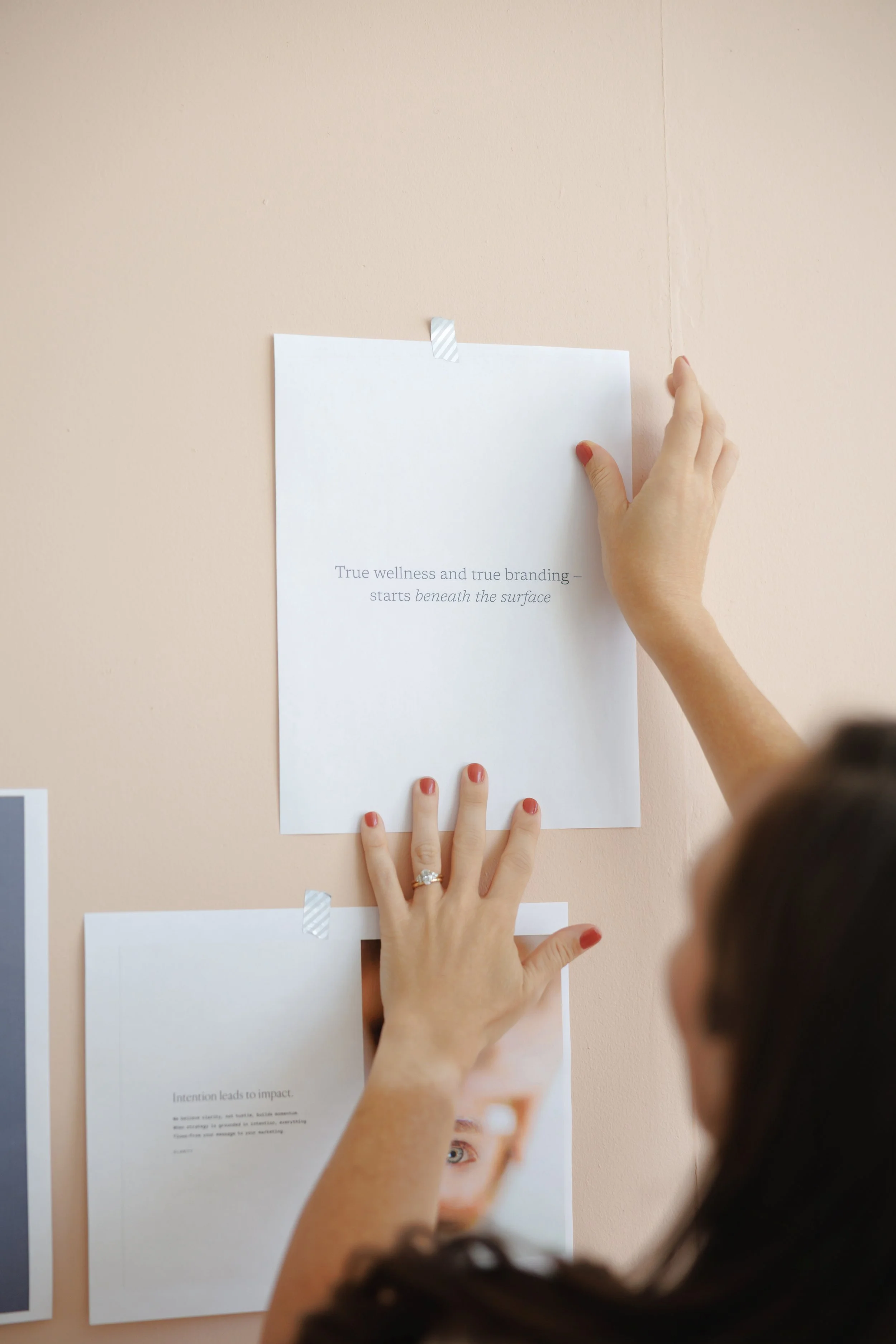 Person taping a printed paper with the text "True wellness and true branding - starts beneath the surface" onto a peach-colored wall.