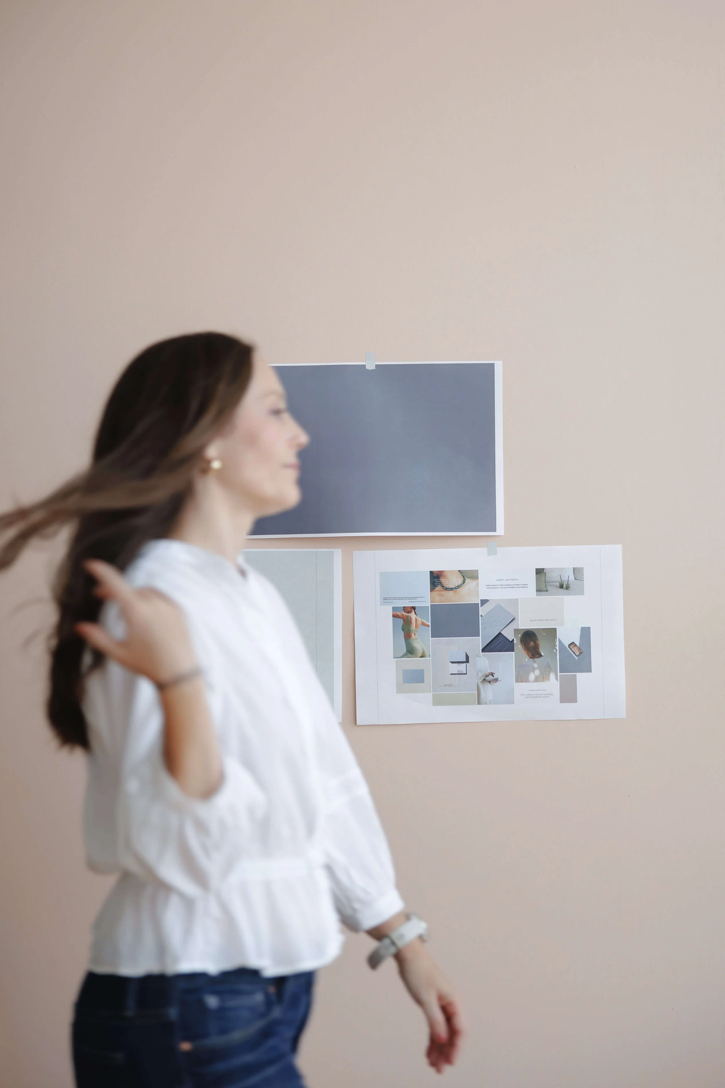 A woman with long brown hair wearing a white blouse and dark jeans stands in front of a beige wall with photographs and a large dark-colored paper pinned to it.