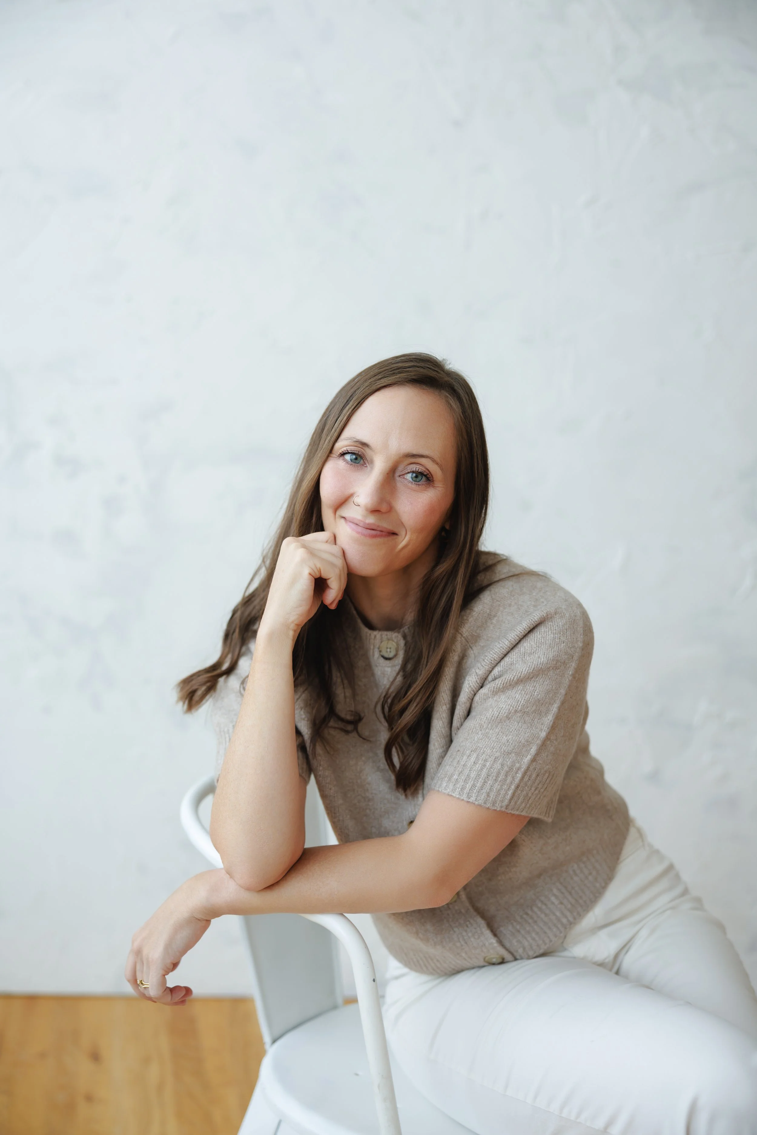 A woman with long brown hair and blue eyes sitting on a white chair. She is resting her chin on her right hand and smiling at the camera. She is wearing a beige short-sleeved sweater and white pants, with a plain light-colored wall in the background.