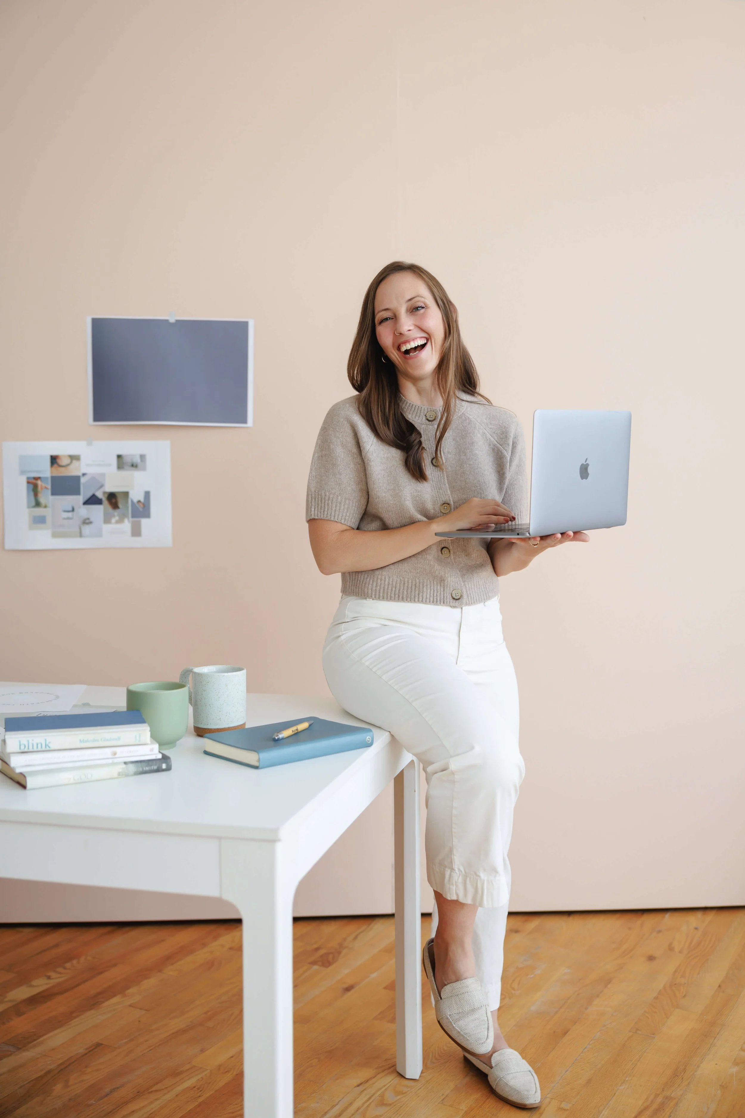 A woman with long brown hair, wearing a beige button-up short-sleeve top and white pants, laughing and holding a silver laptop. She is standing next to a table with books, cups, a notebook, and a pen, in a room with light beige walls and wooden floor.