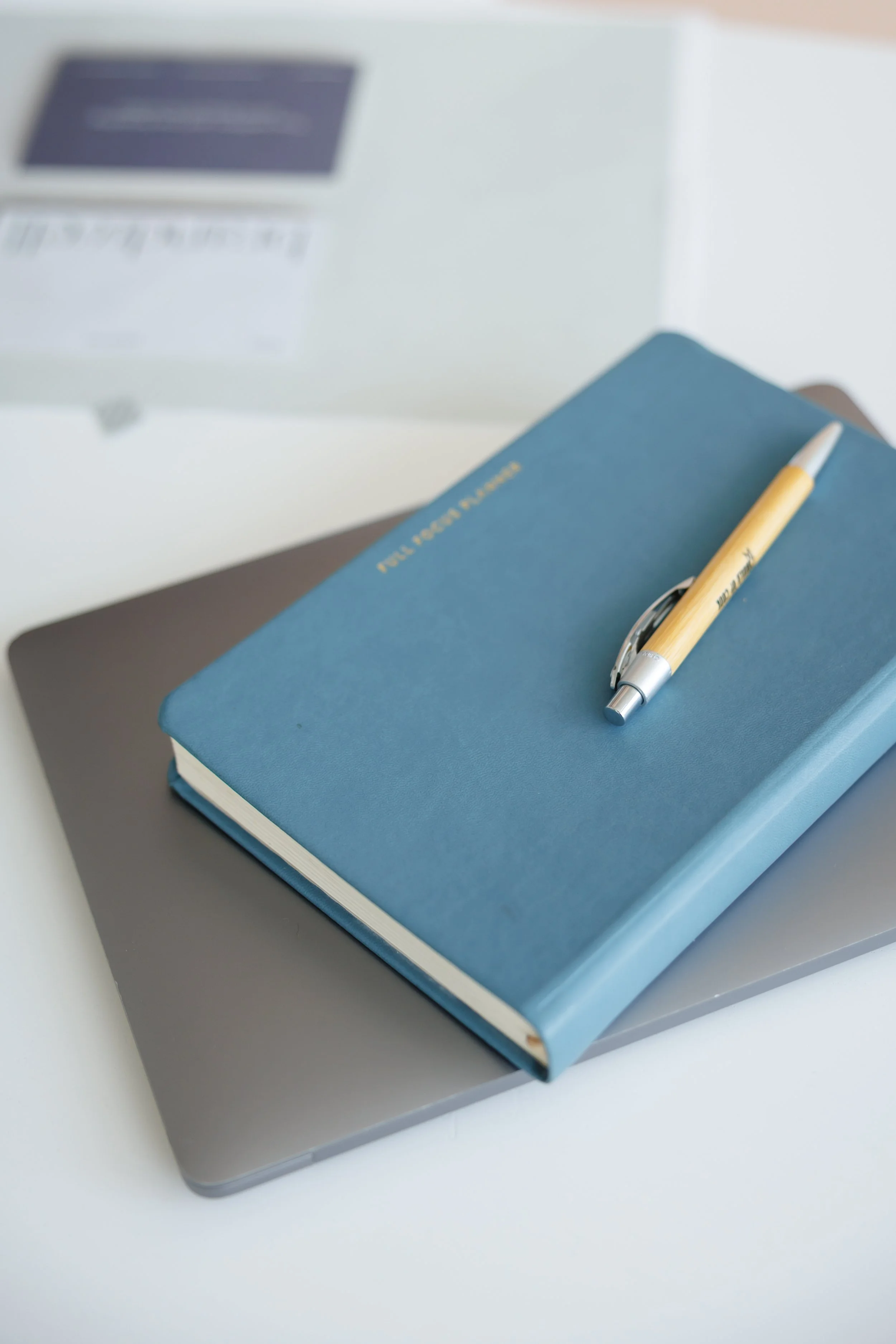 A closed blue planner with 'Put Your Goals Here' written on the cover, a pen on top, a silver laptop underneath, and a white box in the background.