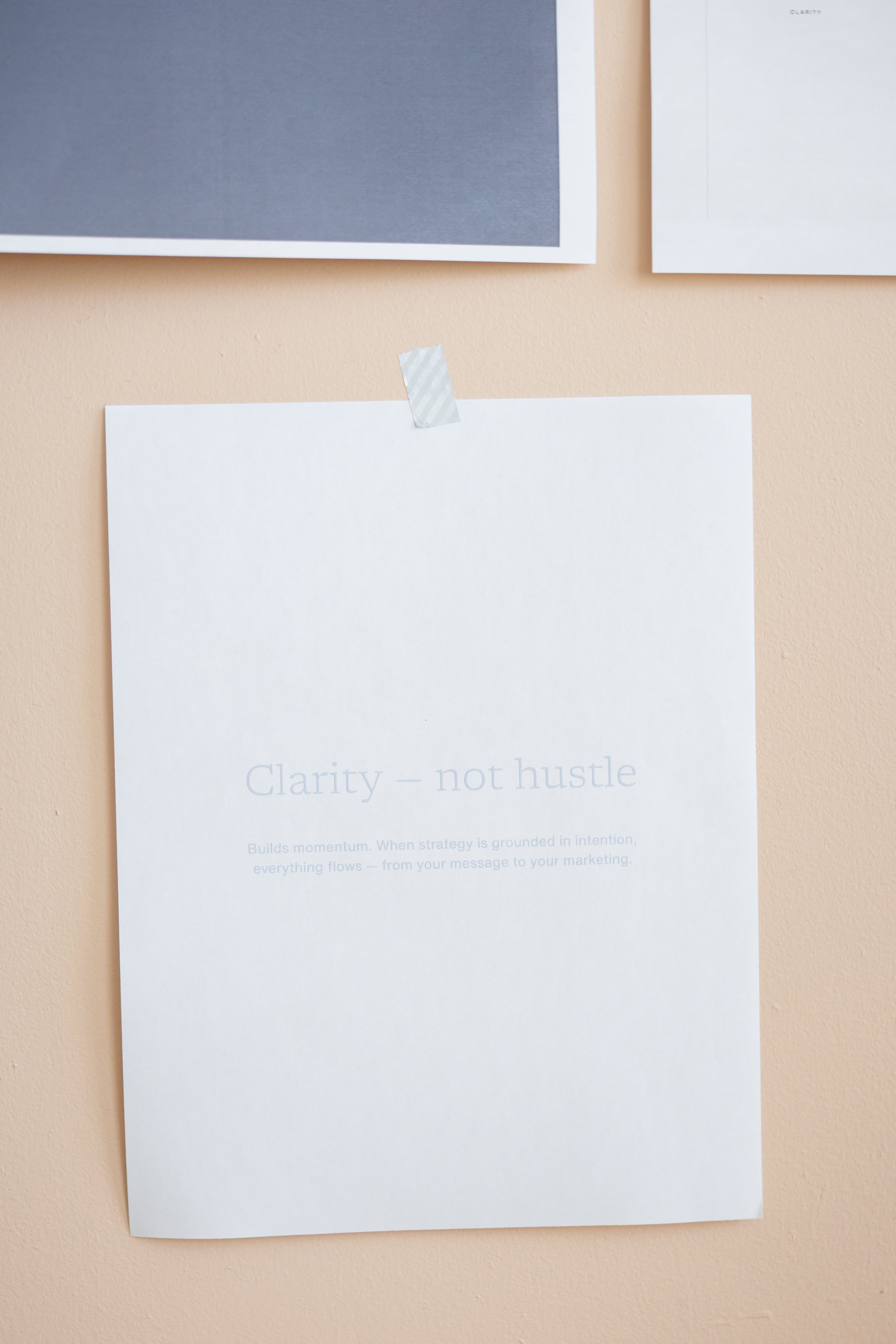 A white sheet of paper taped to a light beige wall, with the text "Clarity - not hustle" and additional smaller text.
