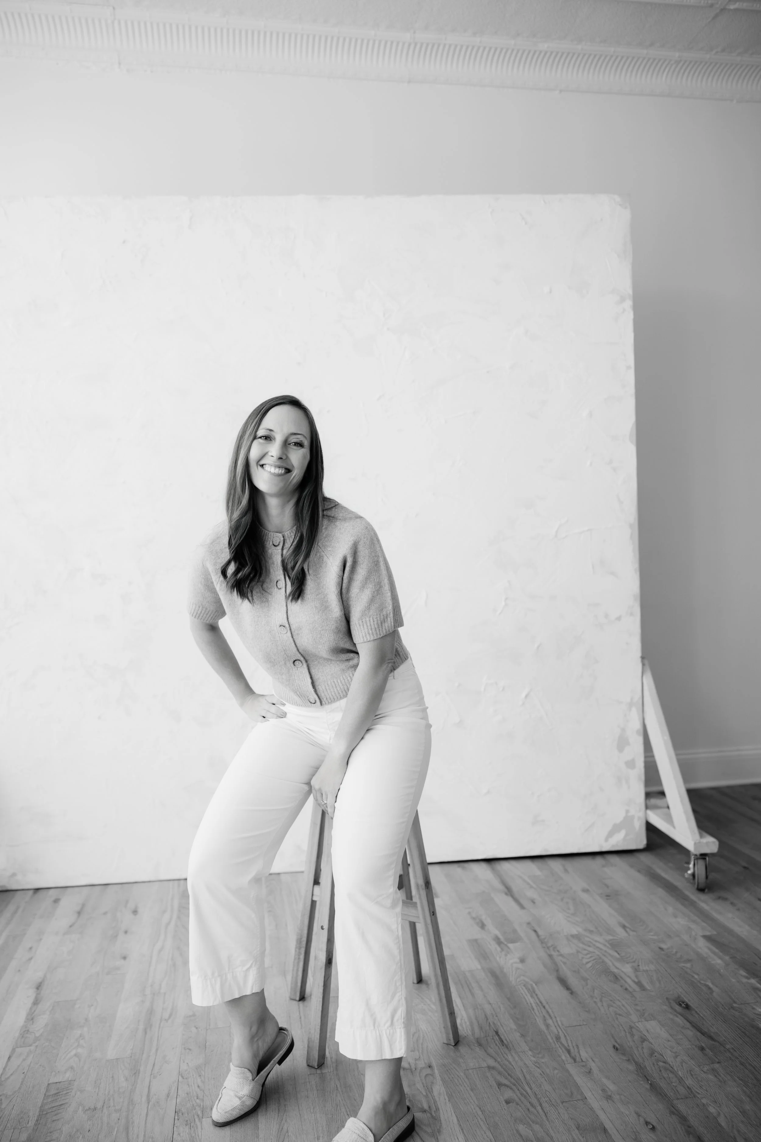 Black and white photo of a woman sitting on a stool, smiling, in front of a large textured white backdrop on a wooden floor.