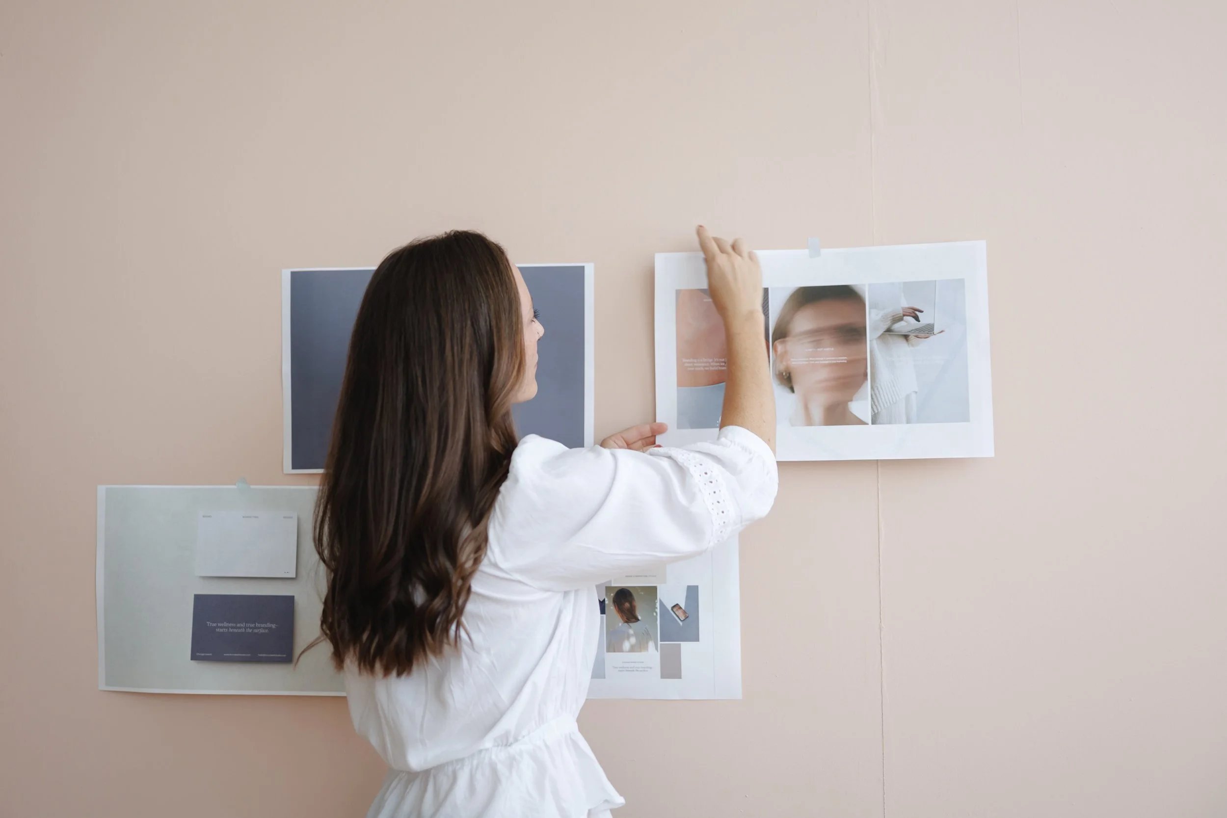 A woman with long brown hair and a white blouse is hanging up or adjusting printed images and documents on a beige wall, with other images and pages already taped or pinned to the wall.
