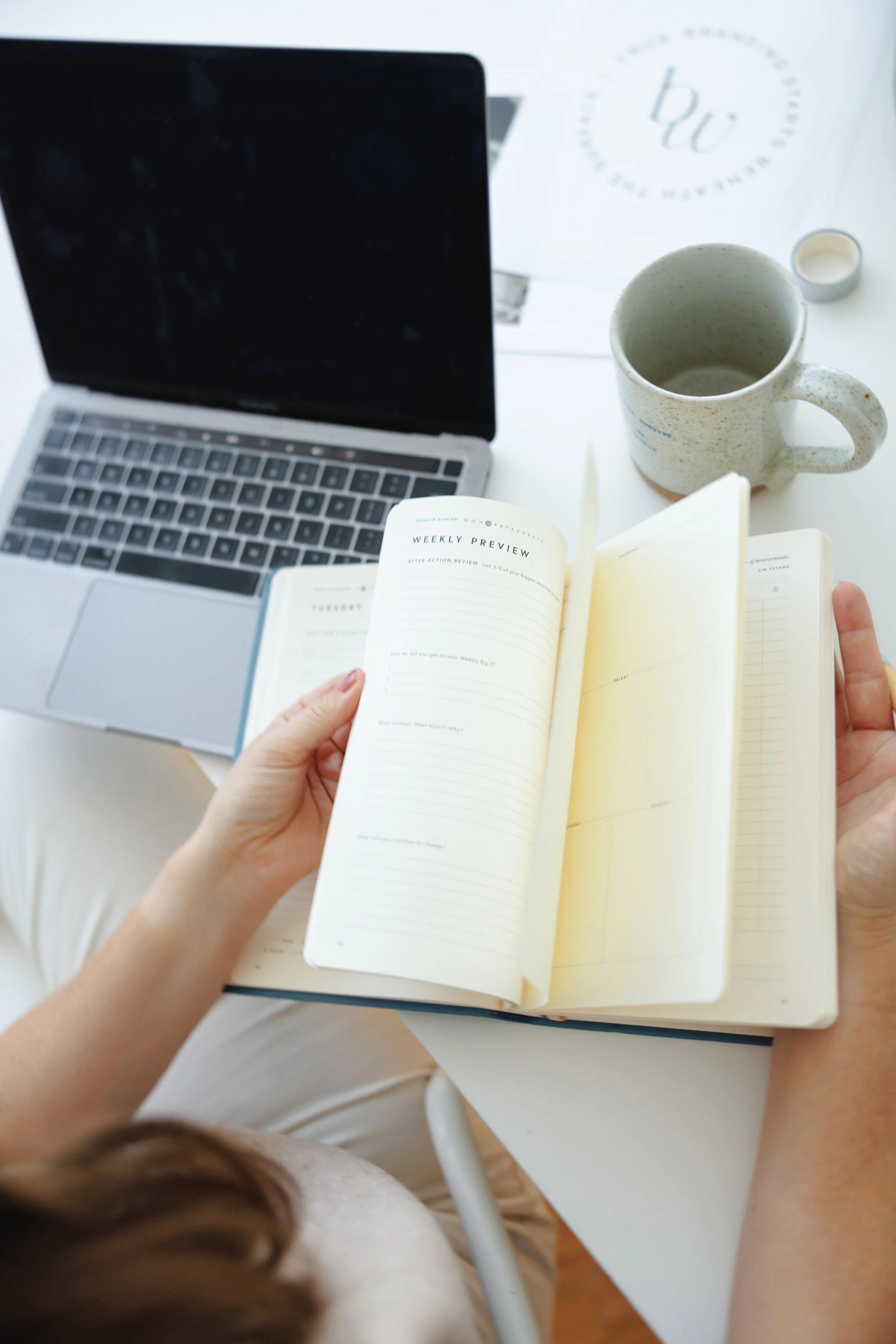 Person holding a weekly planner with a laptop, coffee mug, and other papers on a white desk.