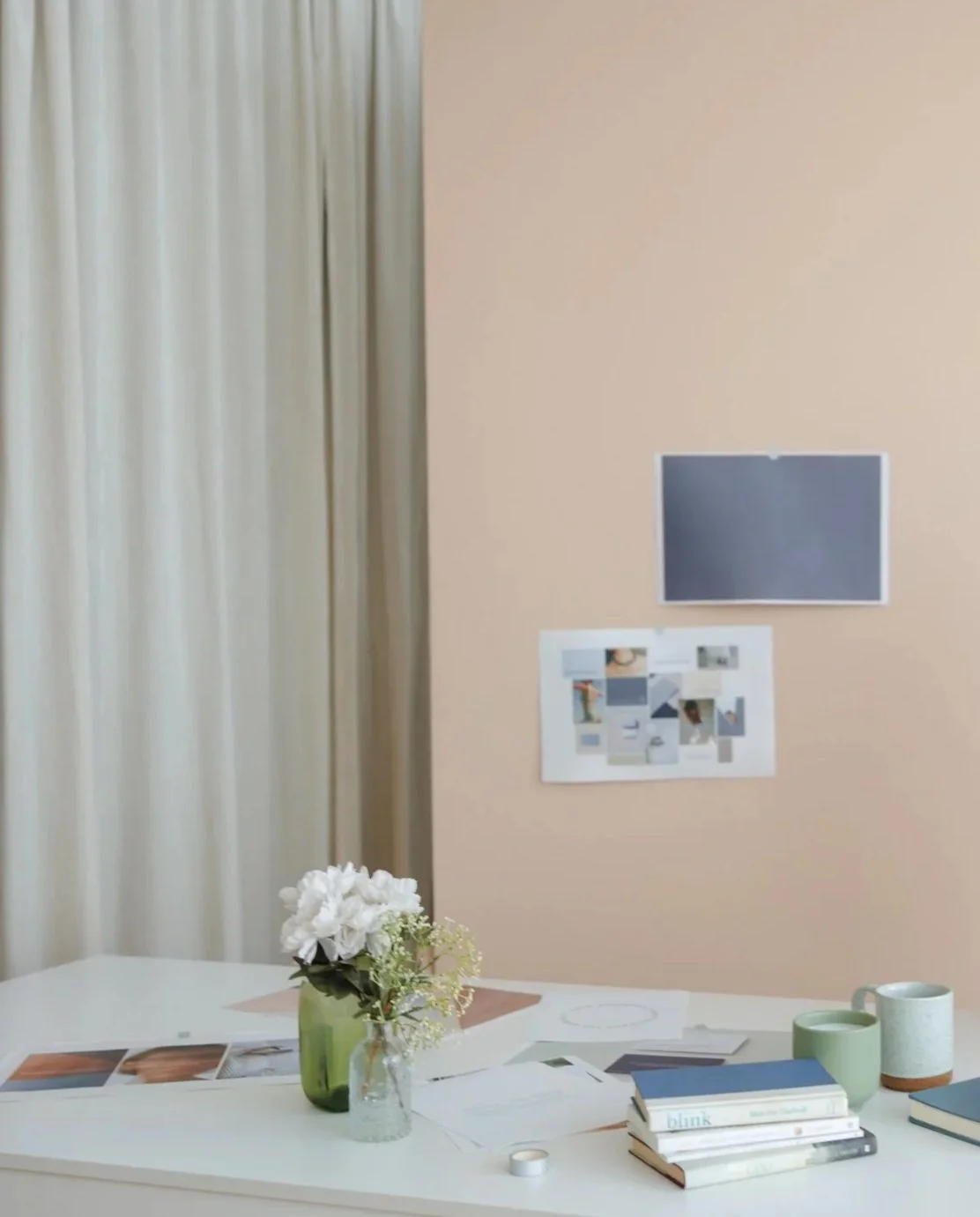 A white table with a green vase containing white flowers, books, notebooks, cups, and papers, set against a beige wall with photos and a dark screen, and cream-colored curtains.