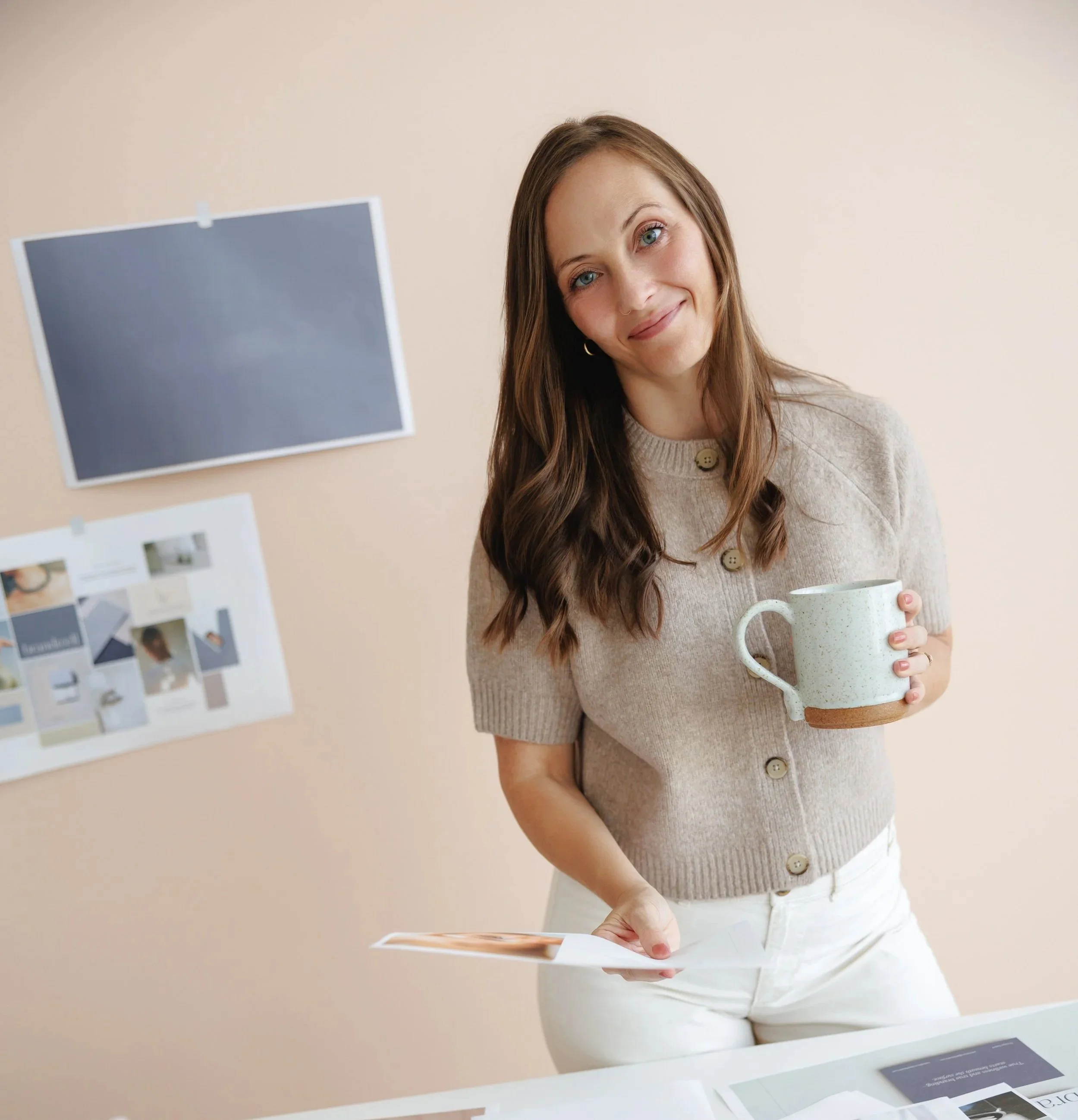 A woman with long brown hair and blue eyes is holding a ceramic mug while standing in front of a beige wall with various interior design swatches and photos displayed.