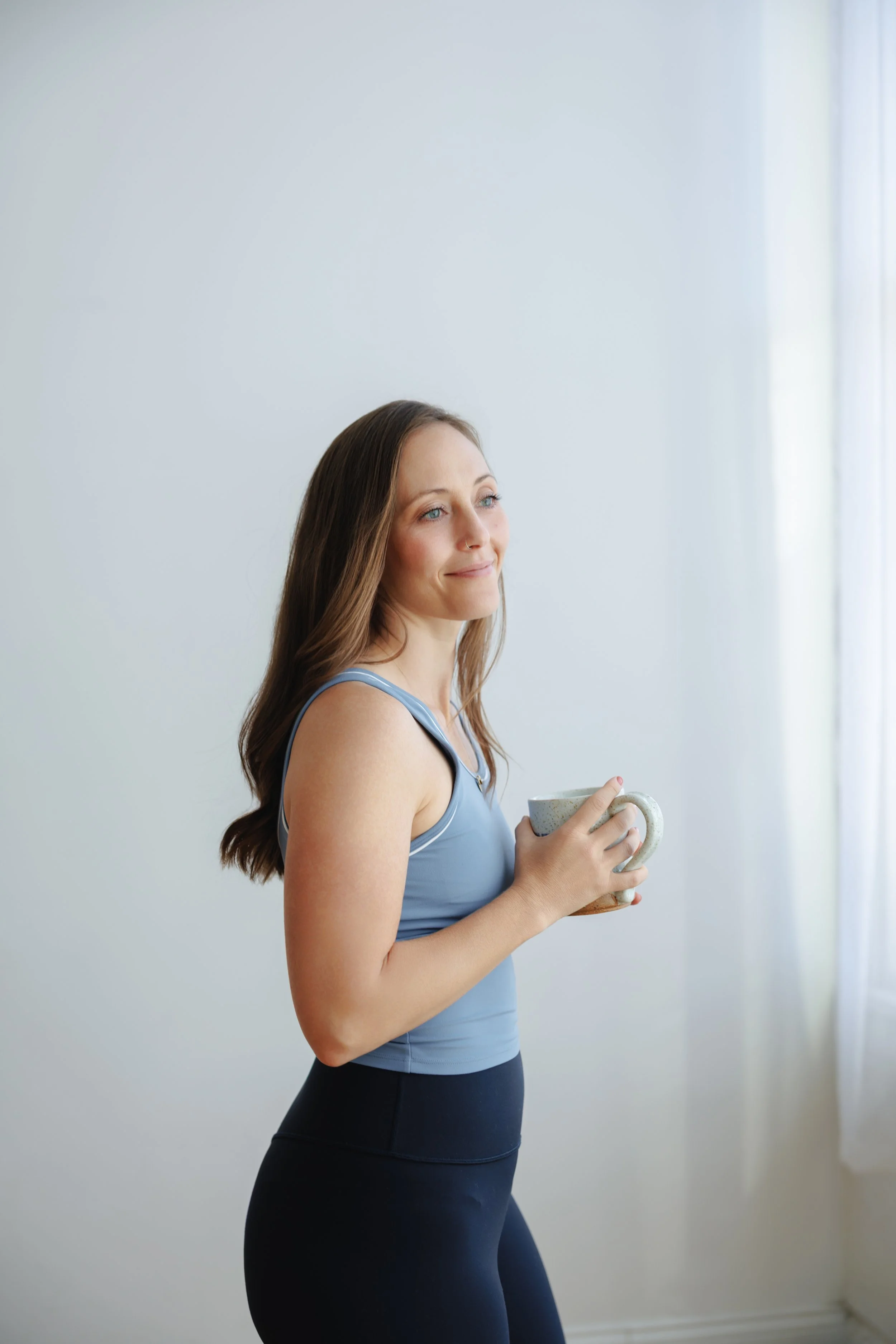 A woman in workout attire holding a mug, standing near a window with light curtains on the right side.