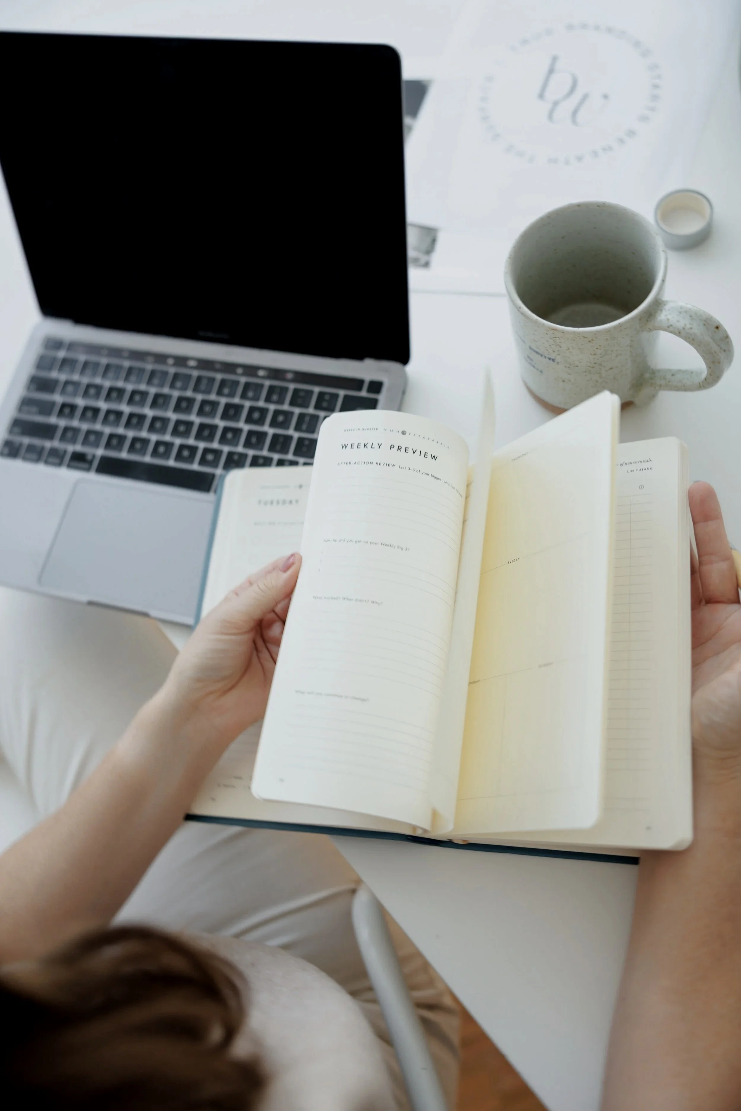 Person holding an open planner over a white table with a laptop, coffee mug, and some papers.