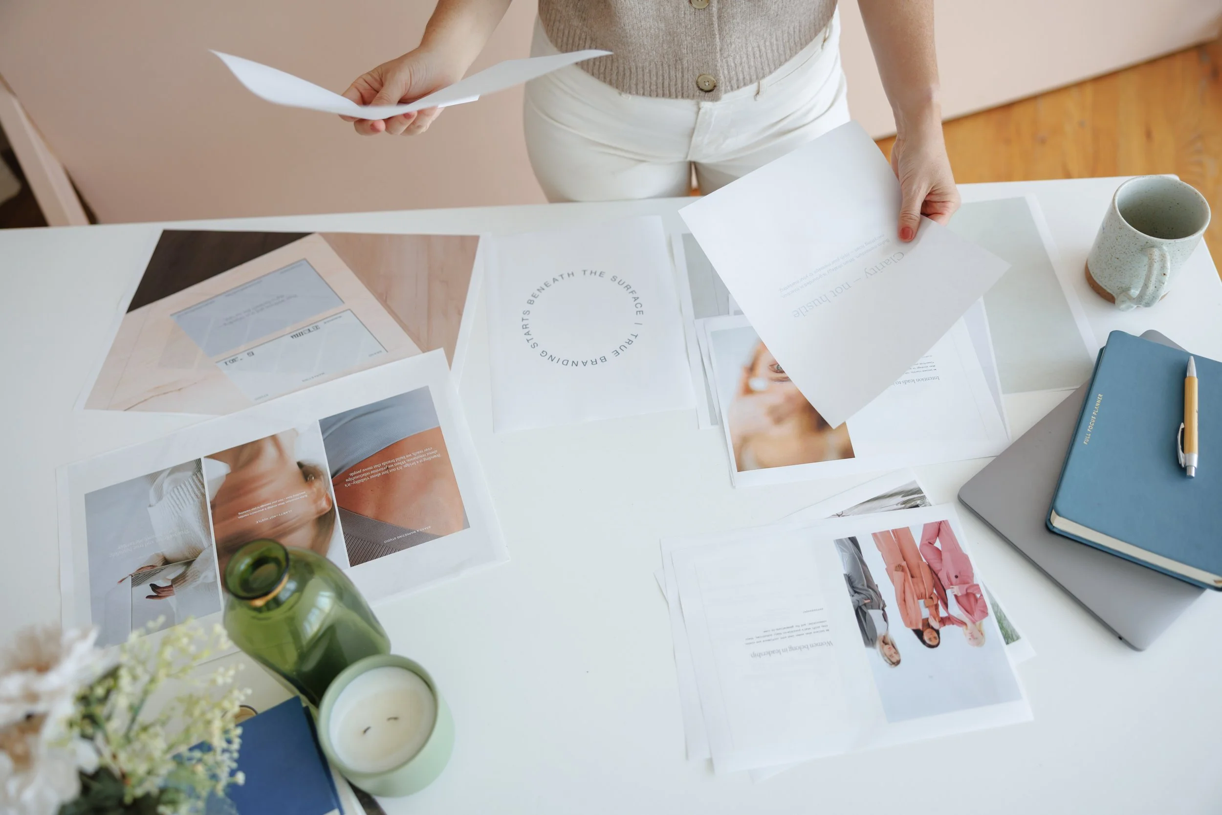 Person standing over a white table with various papers, magazines, a blue notebook, a gold pen, a ceramic mug, a small candle, and a green bottle.